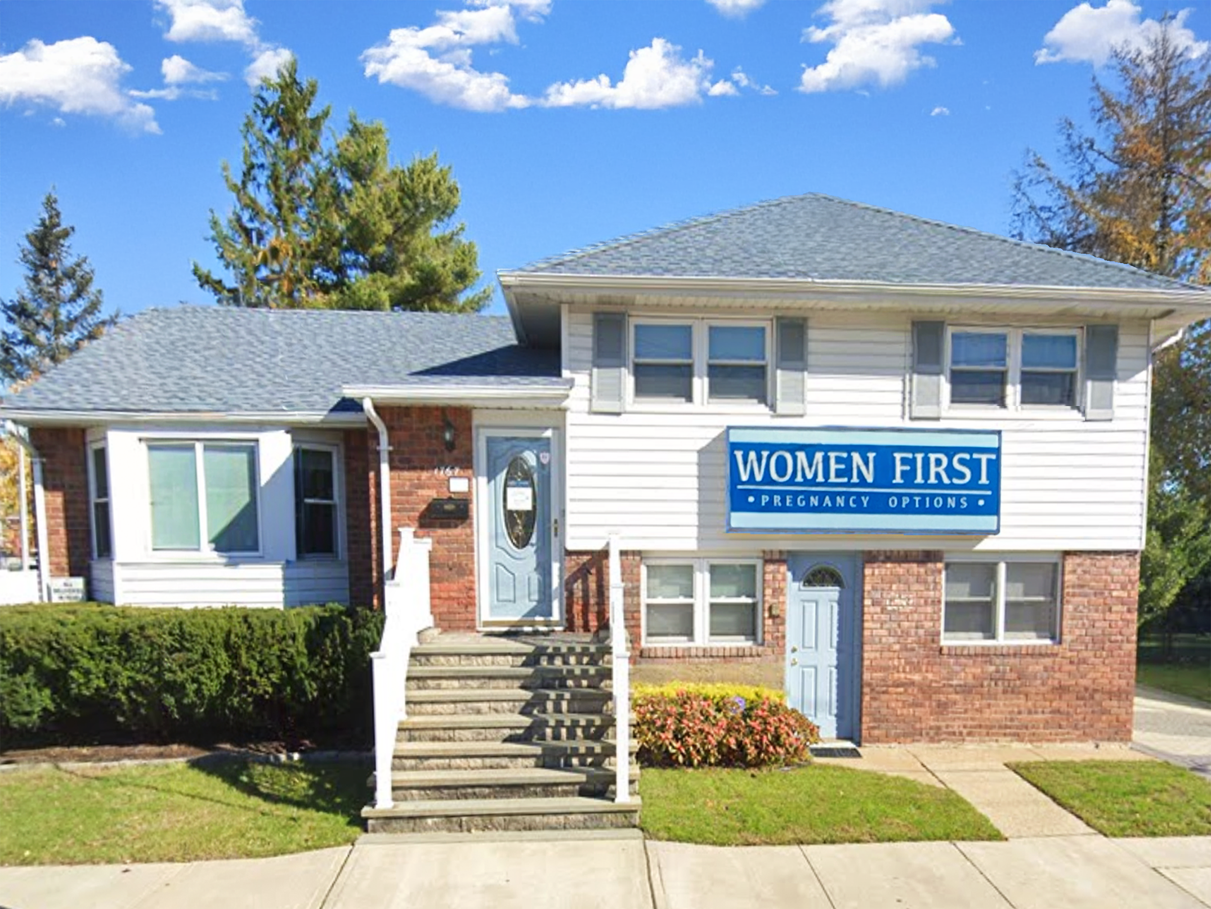 A white residential house with three stories and multiple windows, a front porch with stairs, a lawn with bushes, and a sign that reads 'WOMEN FIRST PREGNANCY OPTIONS' attached to the house. The house has a grey roof, and the background features trees and a clear blue sky.