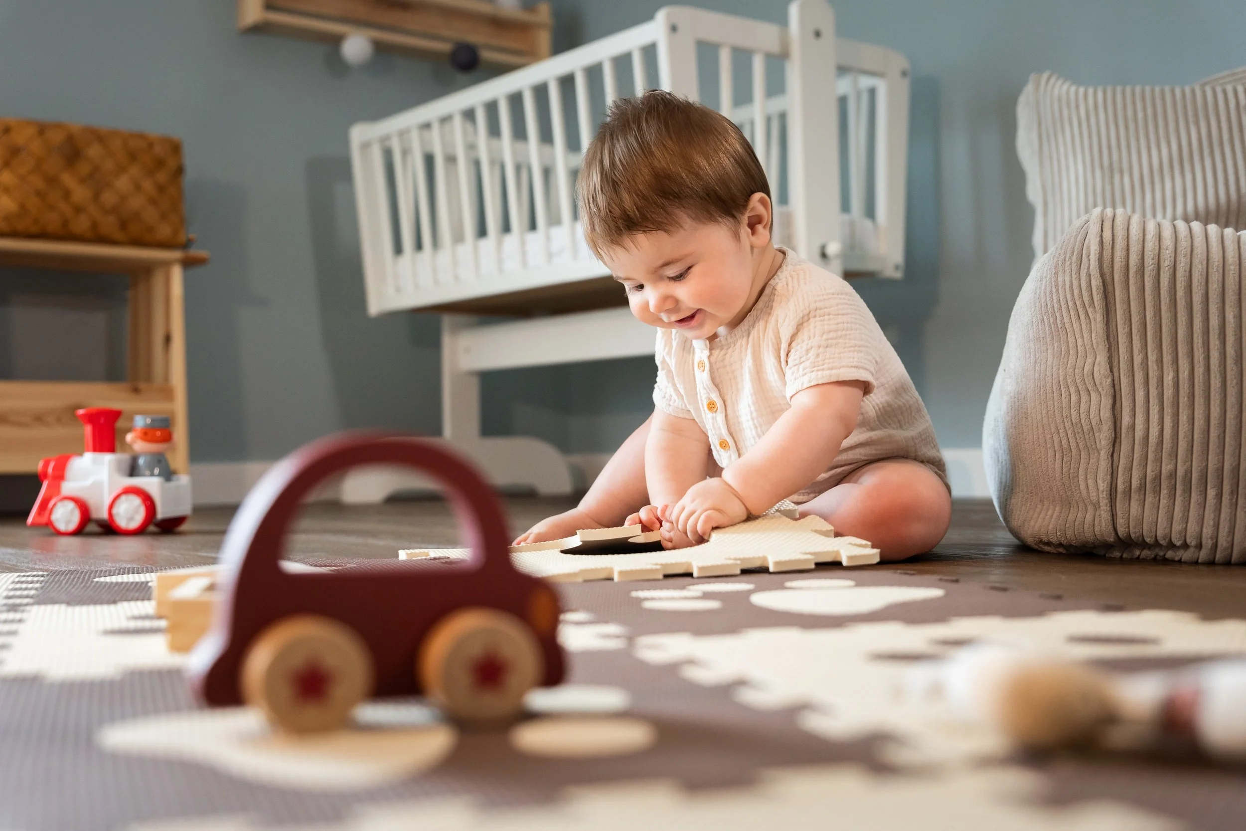 A young child playing on the floor, smiling, with toys around in a room with blue walls and a white crib.
