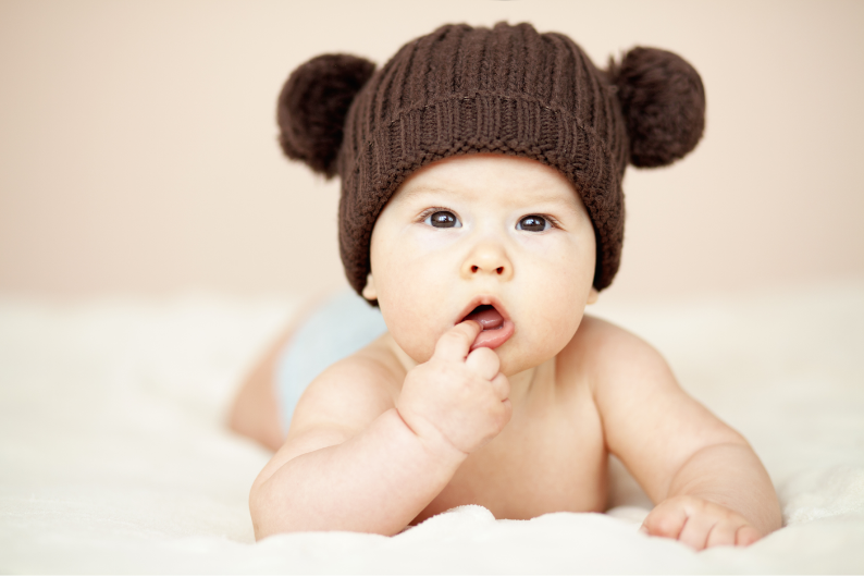 A baby wearing a brown knit hat with bear ears, lying on a white surface, with a curious expression and finger in mouth.