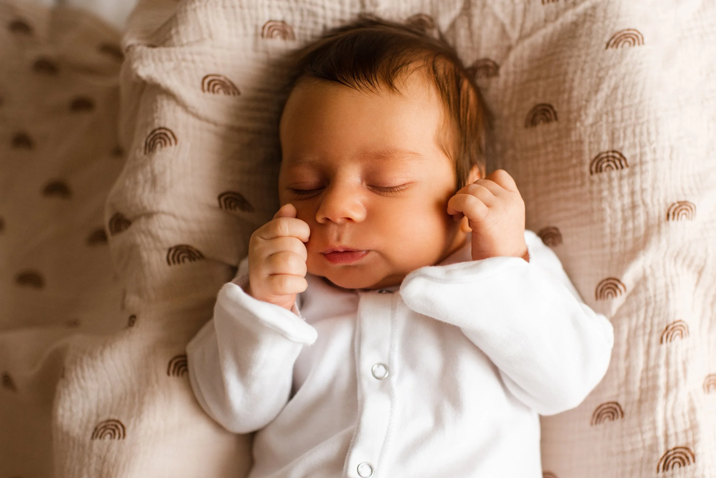 Close-up of a sleeping baby with hands near face, lying on a beige blanket with rainbow prints.