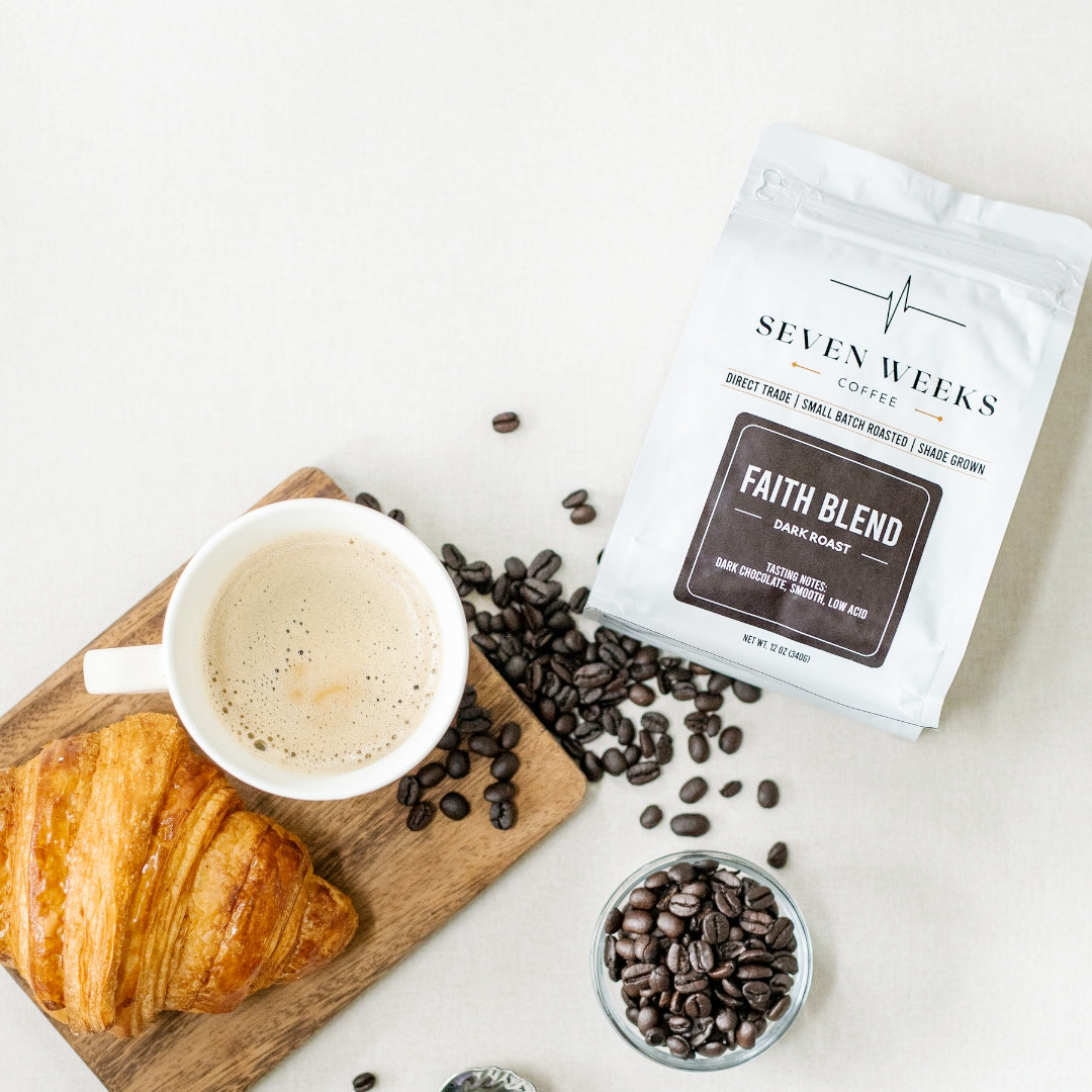 A cup of coffee with a croissant on a wooden tray, coffee beans scattered around, and a bag of Faith Blend dark roast coffee on a white surface.