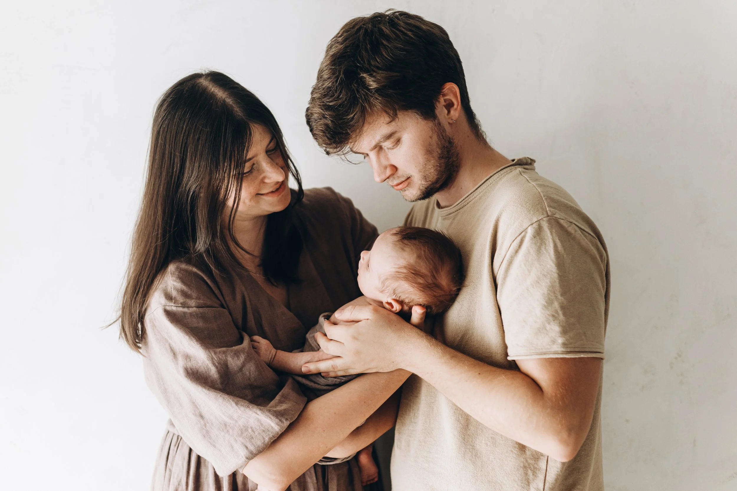 A family holding a newborn baby indoors against a plain wall.