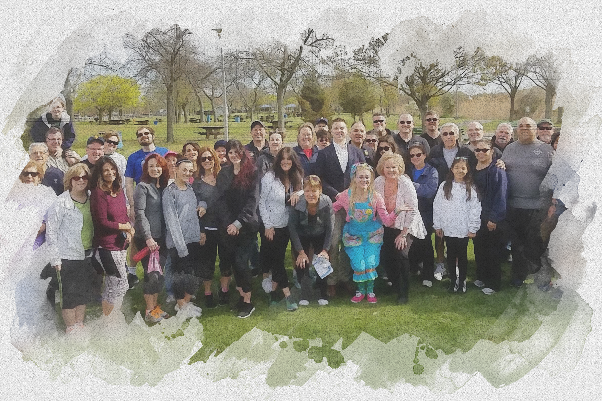 A large group of diverse people, including men, women, and children, gathered outdoors in a park with trees and picnic tables, some wearing sunglasses and casual clothing.