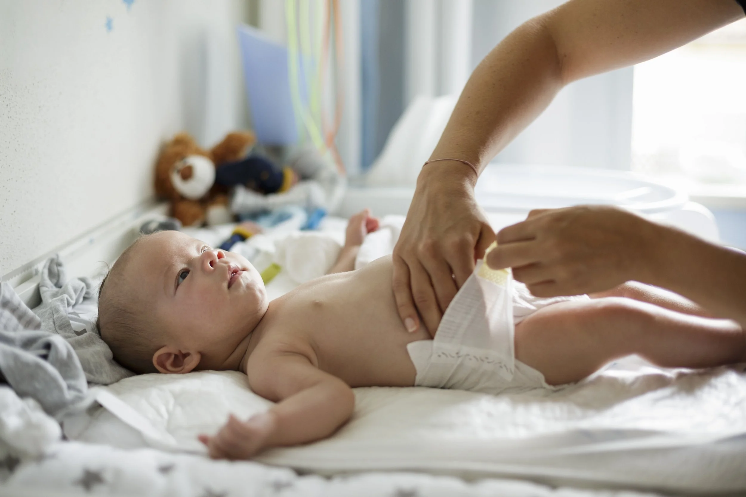 A baby lying on a hospital bed, looking up at an adult who is adjusting or fastening a diaper on the baby.