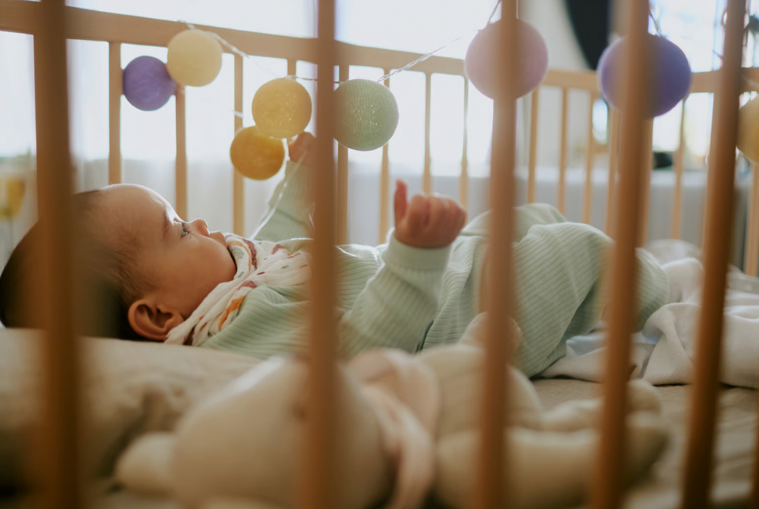 A baby lying in a crib, reaching for pastel-colored hanging decorations, with soft natural lighting.