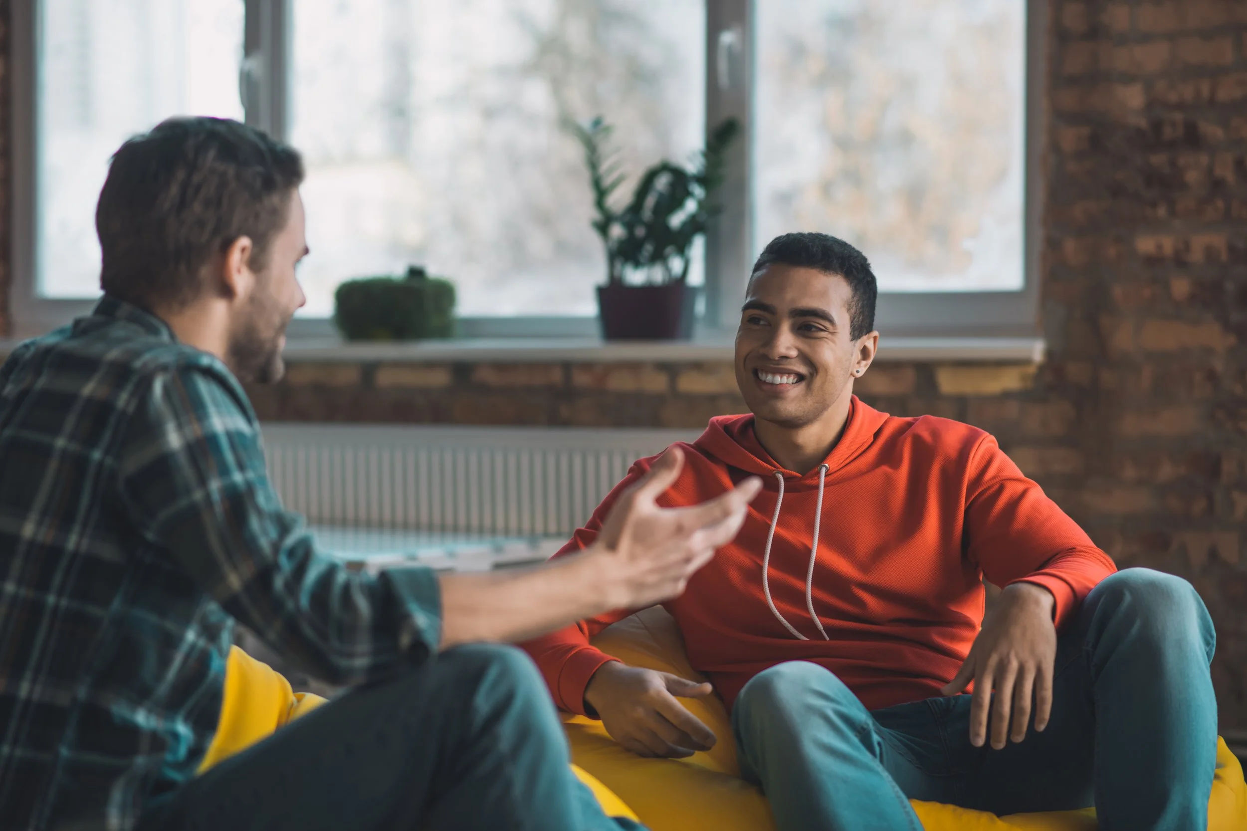 Two young men sitting and talking in a room with large windows and brick walls. One is wearing a plaid shirt, and the other is wearing a red hoodie, smiling during their conversation.