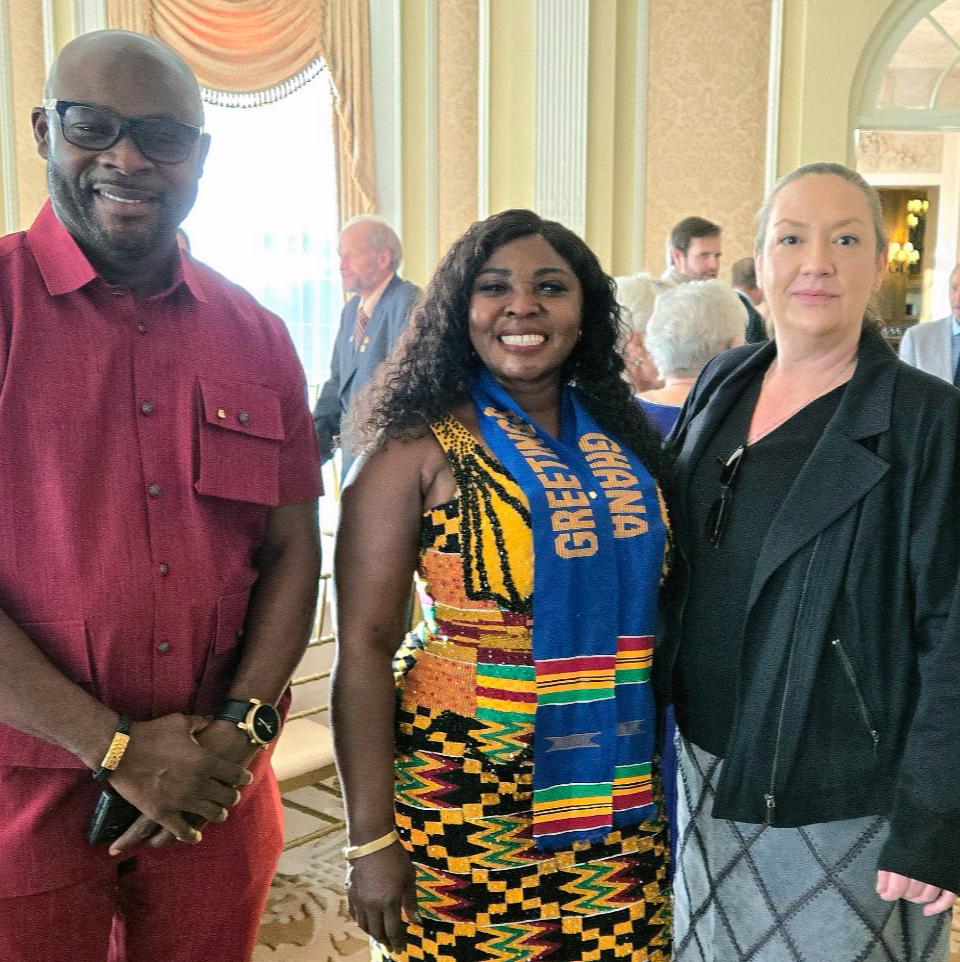 Three people standing together at an indoor event, smiling at the camera. The person on the left is a man wearing a red shirt and glasses, the person in the middle is a woman wearing a colorful dress and a blue Ghana scarf, and the person on the right is a woman wearing a black jacket and a patterned skirt. There are other people in the background engaged in conversation.