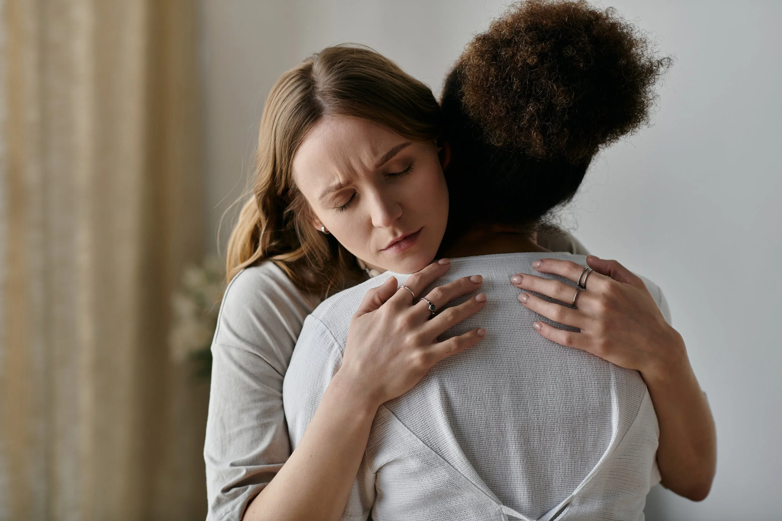 Two women hugging each other tightly, one woman with light skin and brown hair with closed eyes, the other with dark skin and curly hair, both showing a comforting embrace.