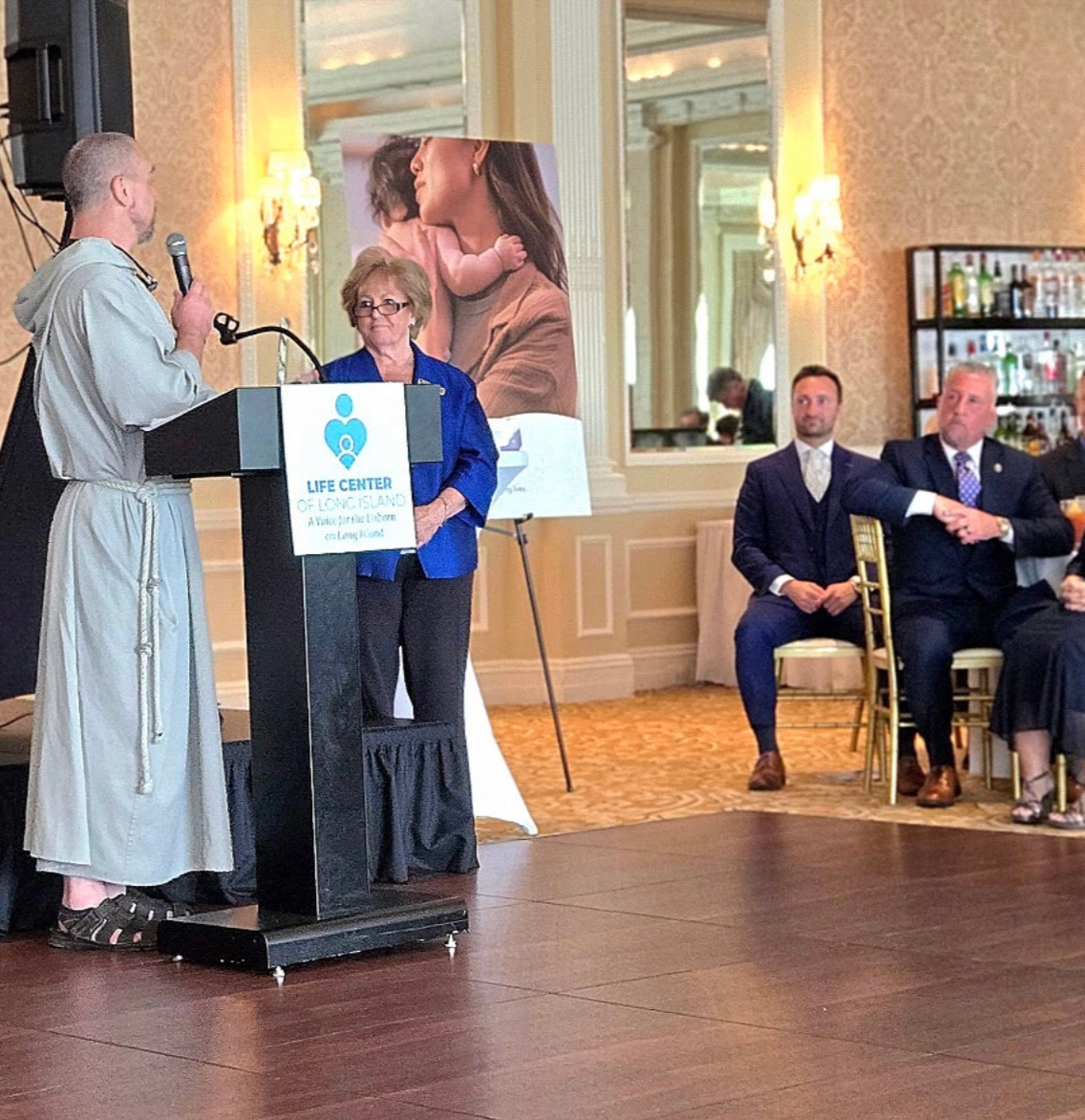 A man in a robe speaking at a podium with a microphone, with an older woman in a blue blazer standing beside him. Several seated attendees are watching, with some wearing suits. The podium has a sign that reads "LIFE CENTER OF LONG ISLAND".