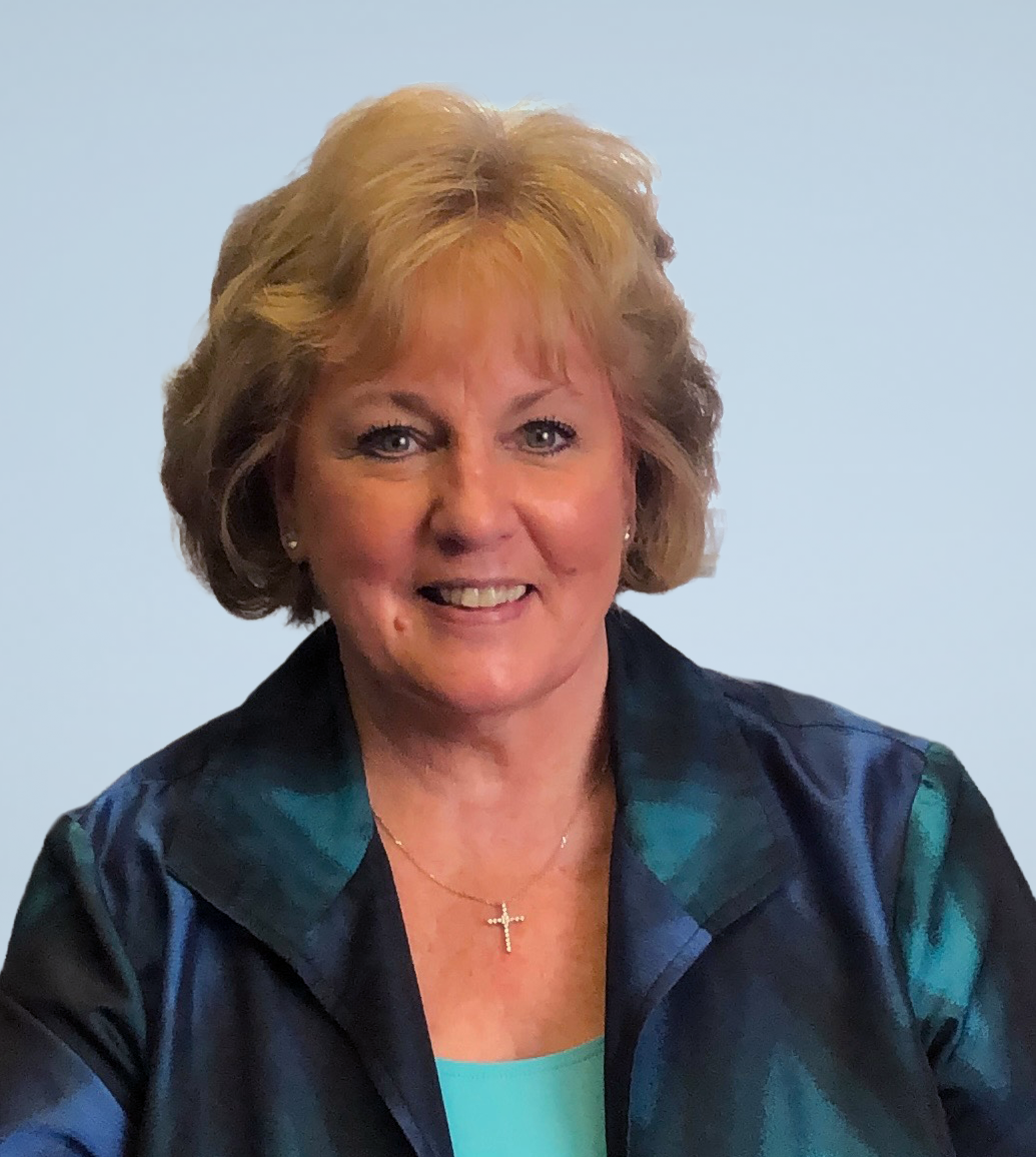 A smiling middle-aged woman with short, blond hair, wearing a cross necklace, a blue top, and a dark blazer, posing against a plain background.