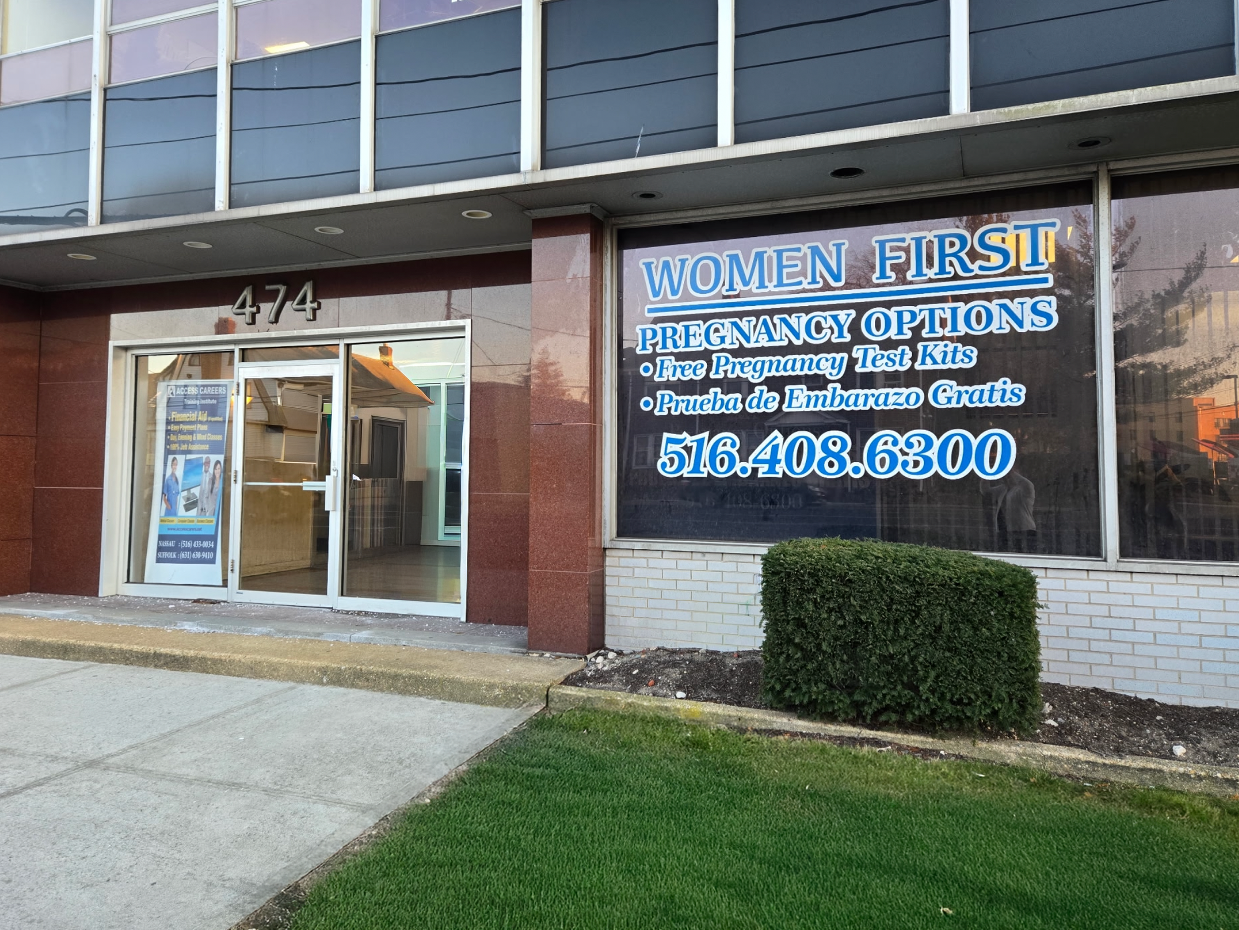 Storefront with large business sign that reads "Women First Pregnancy Options" with additional details in blue text, and the address 474 on the building. The storefront has glass doors and windows, with a small shrub in front and a well-maintained grassy area.