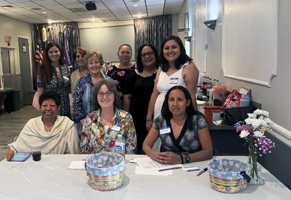 Group of women gathered around a table in a meeting or event room, some sitting and some standing, with a table decorated with flowers and baskets in front of them.