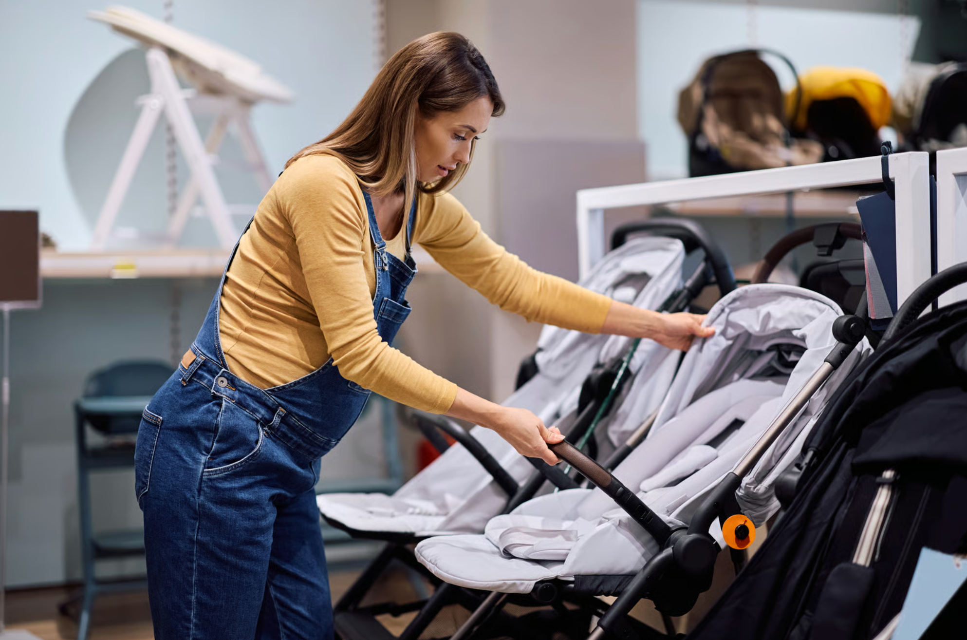 A woman wearing a yellow long sleeve shirt and blue overalls is arranging strollers in a store or showroom.
