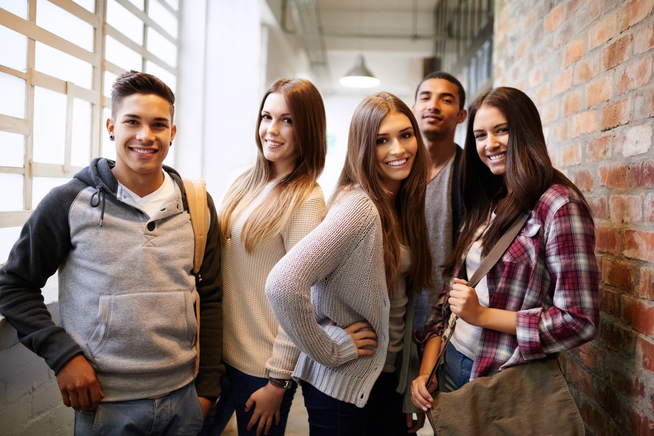 Group of five diverse young adults standing together indoors, smiling, against a brick wall and large window.