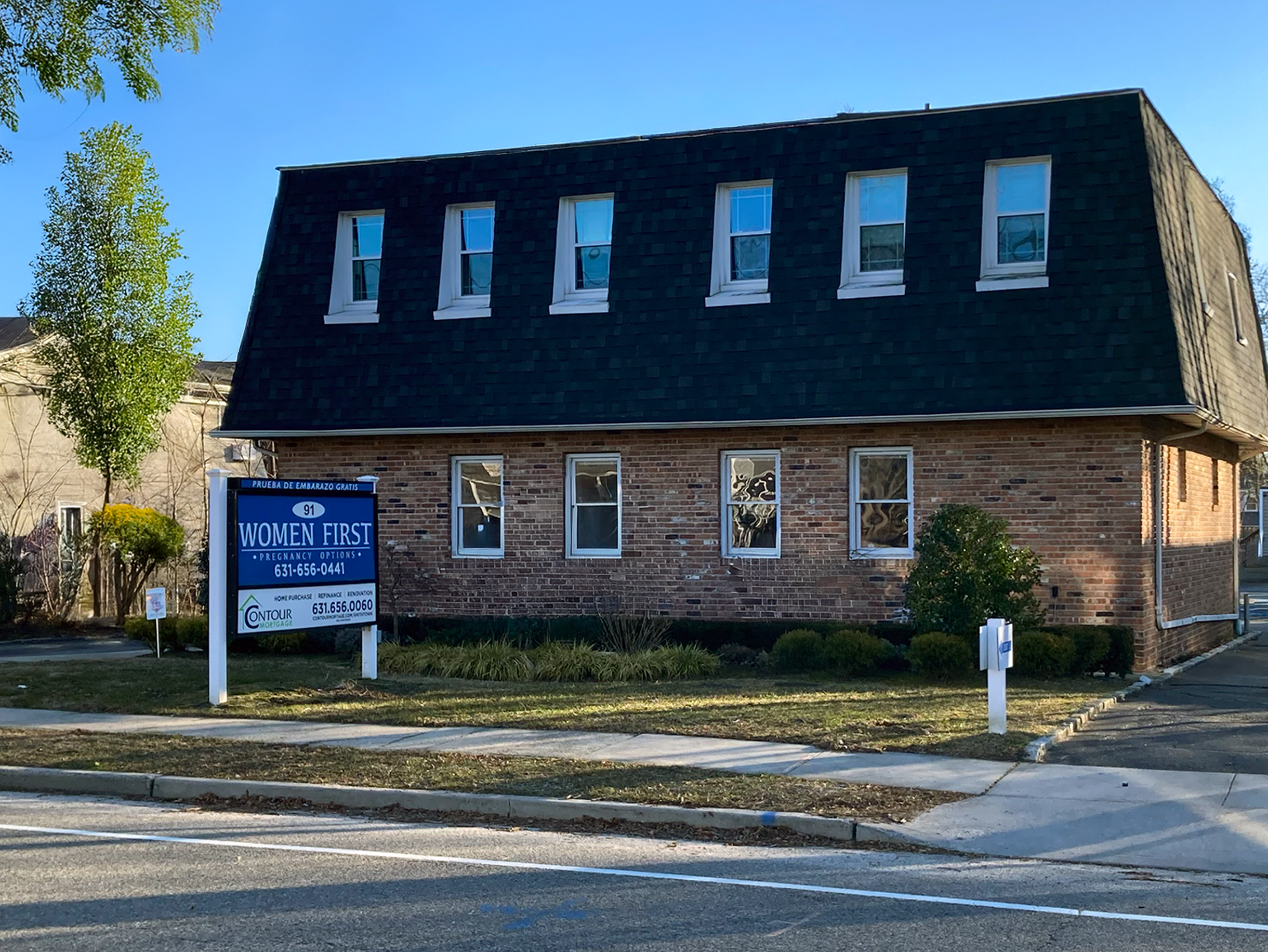 A brick building with a dark roof and multiple windows, some of which have air conditioning units, standing against a clear blue sky. There are trees to the left and a sign in front advertising women's health services.