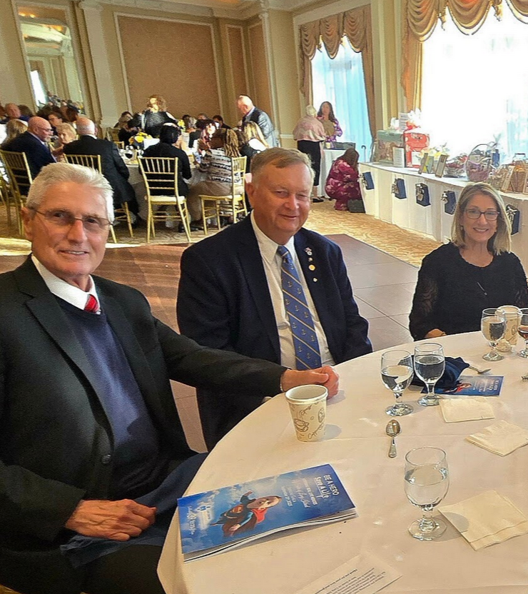 People seated at a round table during a formal event in a decorated banquet room, with more guests seated and a table with gift bags and decorations in the background.