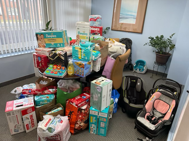 Room filled with baby diapers, wipes, and baby supplies on a table and on the floor, along with a baby car seat, a stroller, and a large potted plant in the corner.