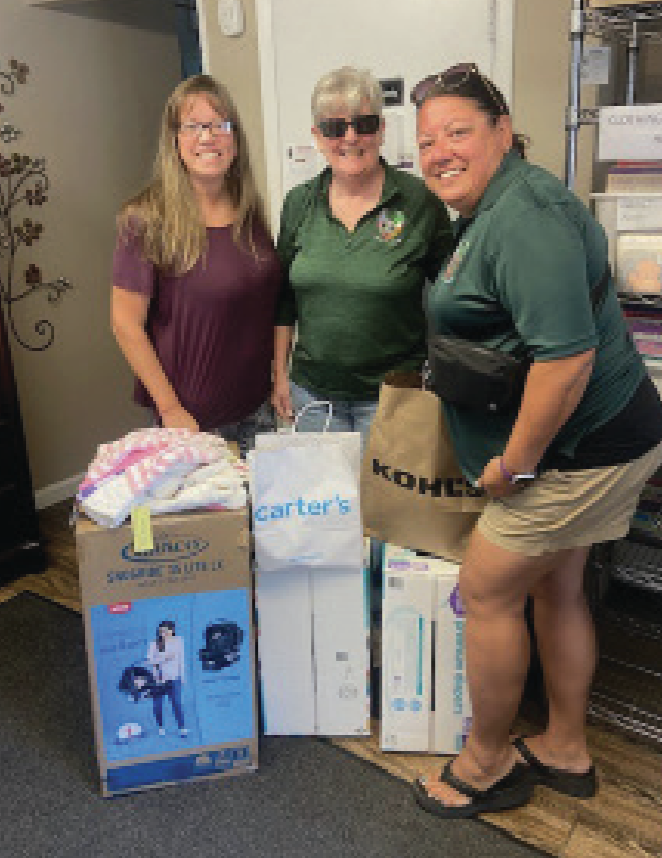 Three women standing behind a collection of donated items, including a baby crib, bags, and boxes, inside a room with shelves and a wall decal.