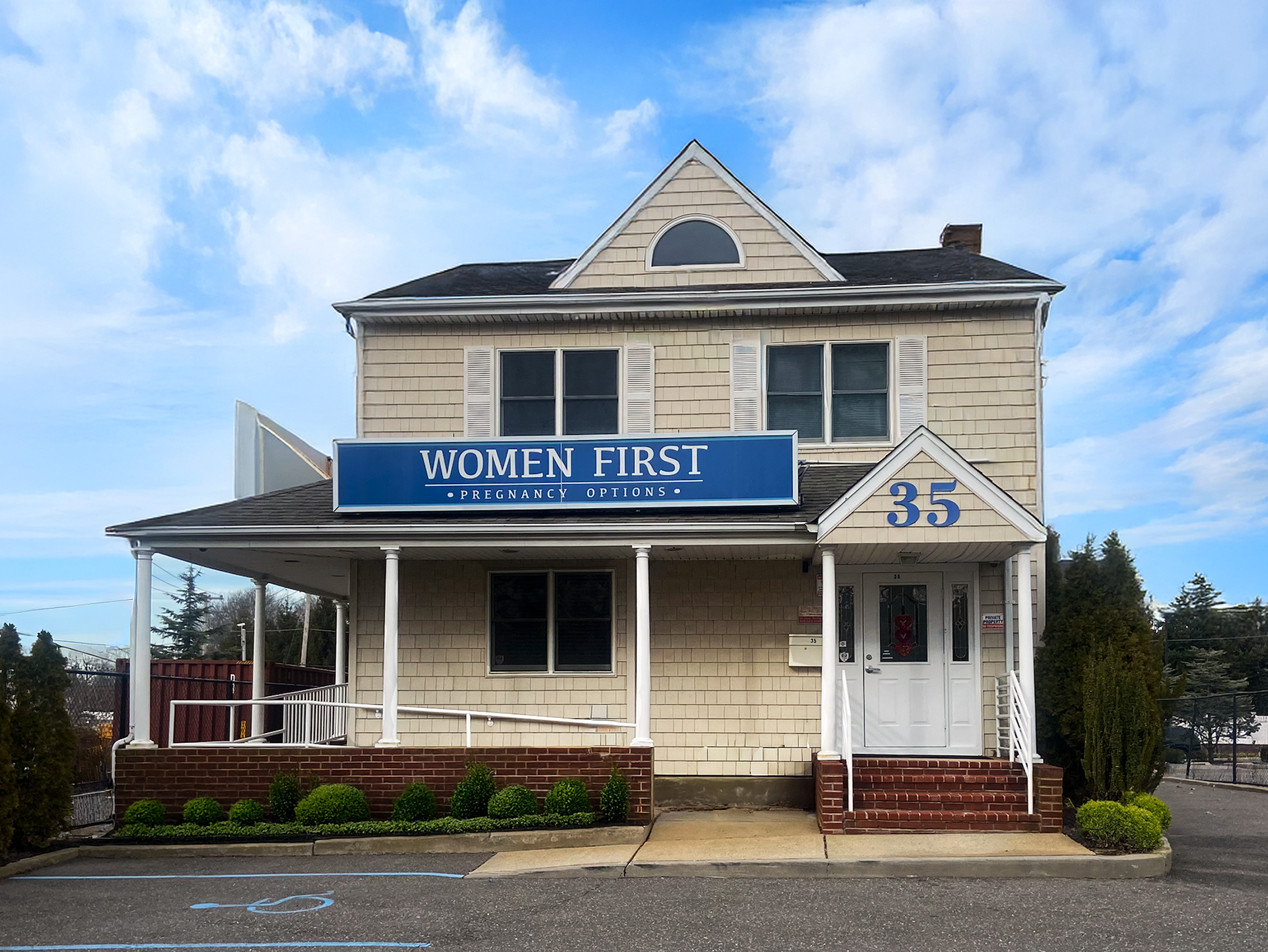 A two-story beige house with white trim, a porch supported by white columns, and a sign that reads "Women First Pregnancy Options." The house has a gabled roof with a semi-circular window, several shutters on the windows, and a small garden with green bushes in front. There is a parking area with blue handicap markings in the foreground, and the sky is partly cloudy with patches of blue.