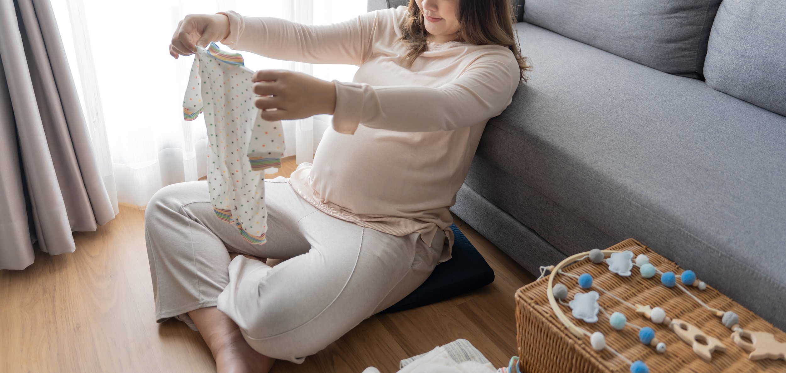 A pregnant woman sitting on the floor, holding a pair of baby socks with colorful dots, near a sofa and wicker basket with blue and white felt balls and baby toys.
