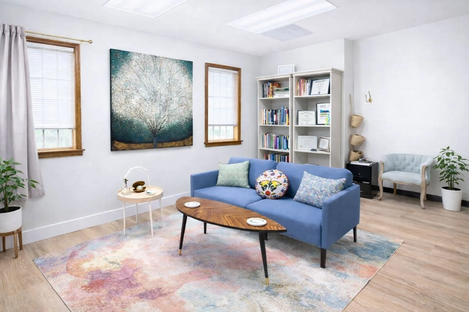 Living room with light wood flooring, a pastel-colored rug, a blue sofa with decorative pillows, a wooden coffee table, a white bookshelf with books and certificates, a small round side table with a lamp and decorative items, a white armchair, green potted plants, two windows with white blinds and curtains, and artwork on the wall.