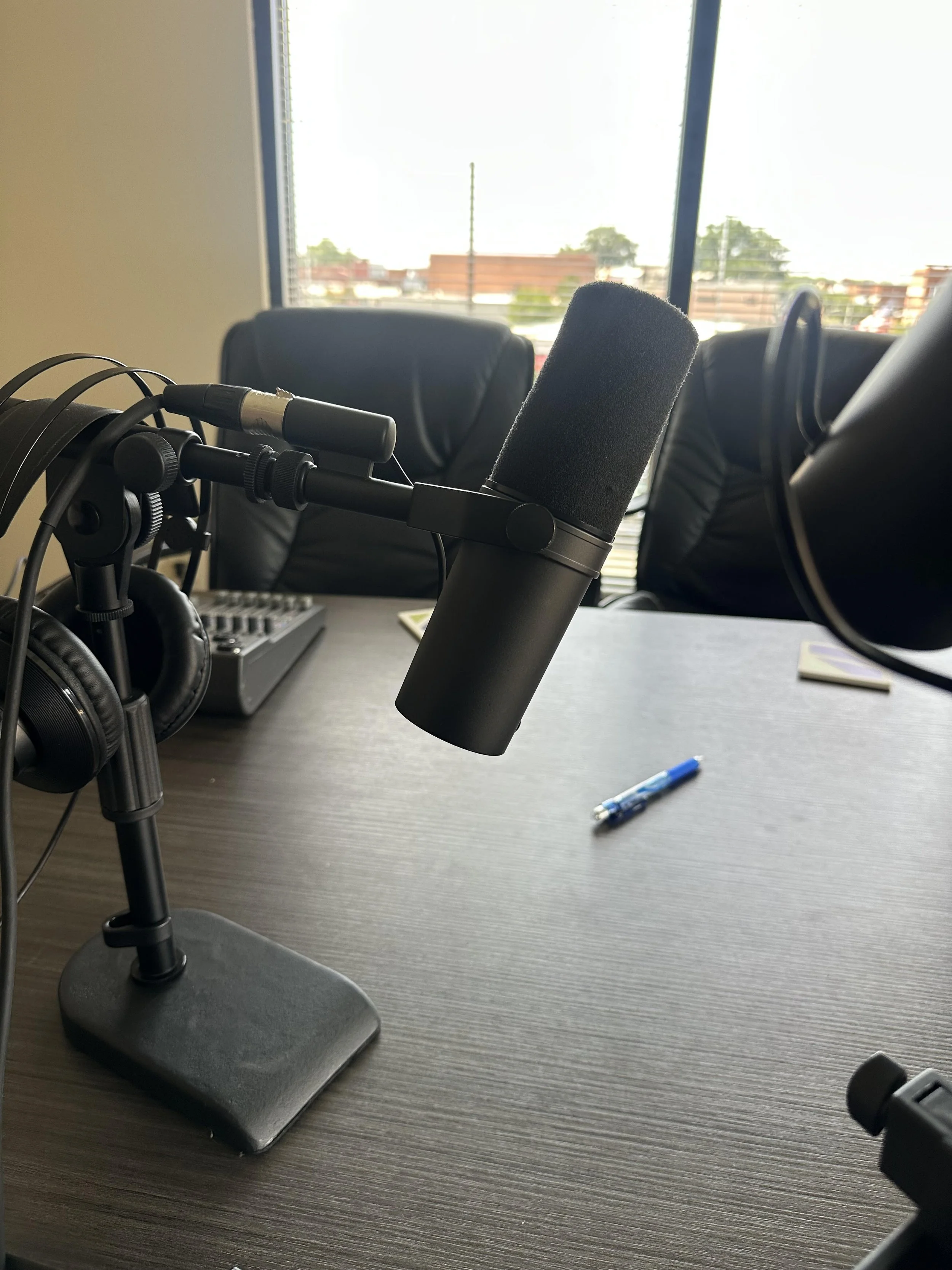 Podcast recording setup on a conference table with a microphone, headphones, a pen, and a sound mixer, in a room with windows and chairs.