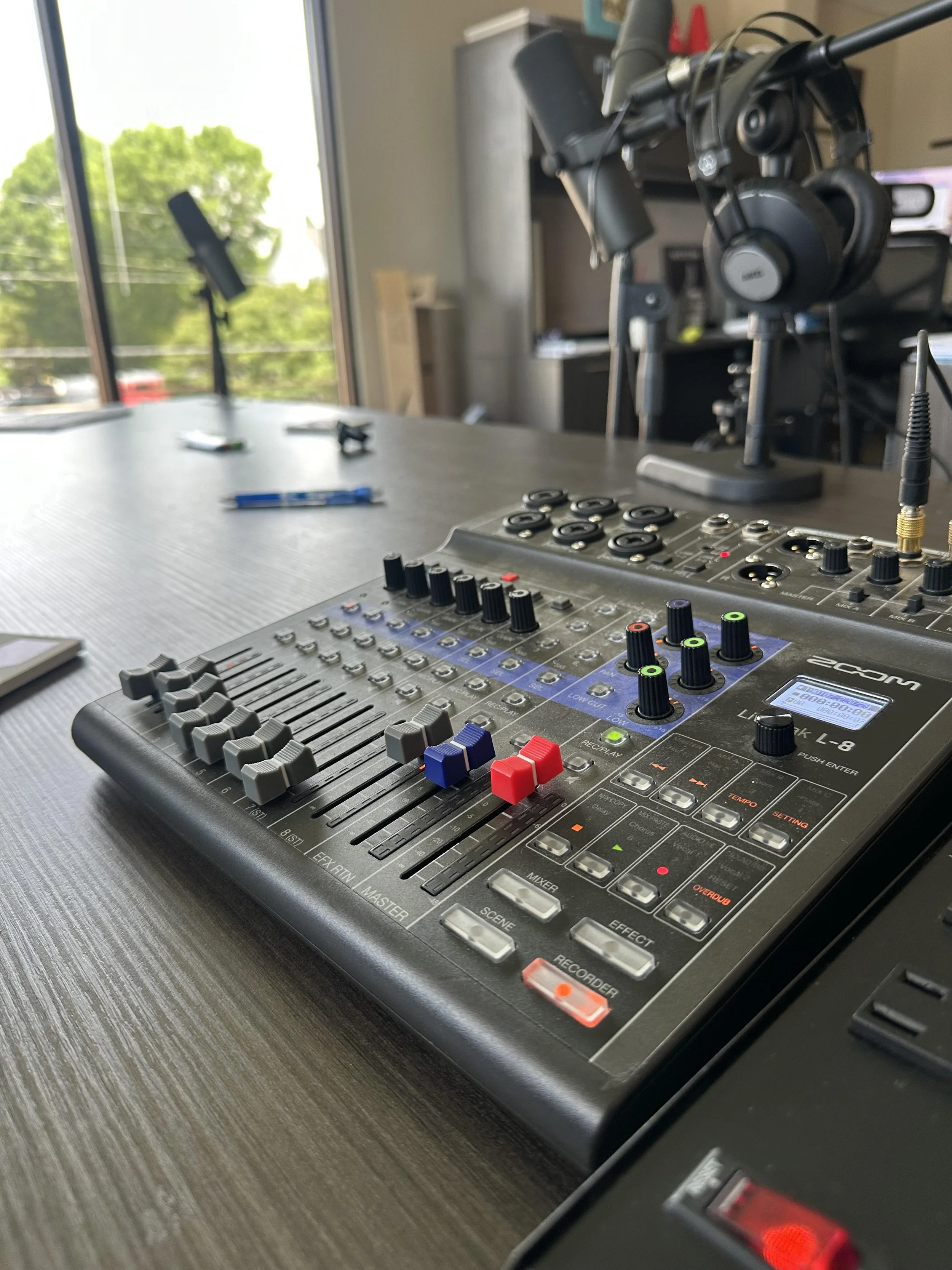 A music production studio featuring a digital audio mixer with various faders and knobs, a microphone on a stand, and headphones hanging on a rack, with natural light coming through large windows showing green trees outside.