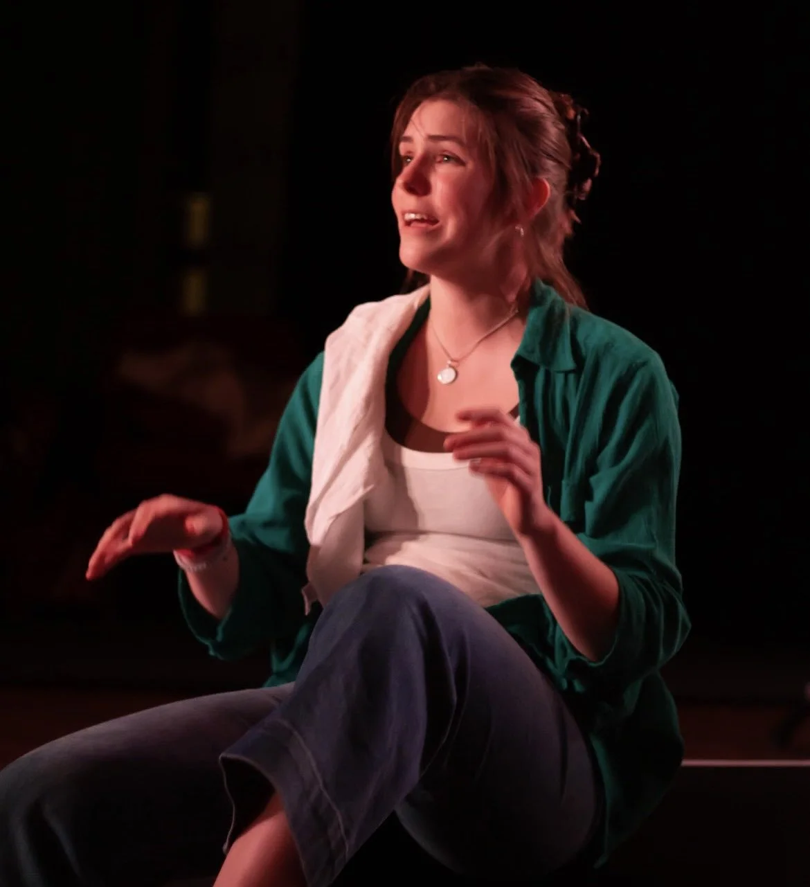 A young woman sitting with a distressed expression, wearing a green shirt over a white top, with a towel draped over her shoulder, against a dark background.