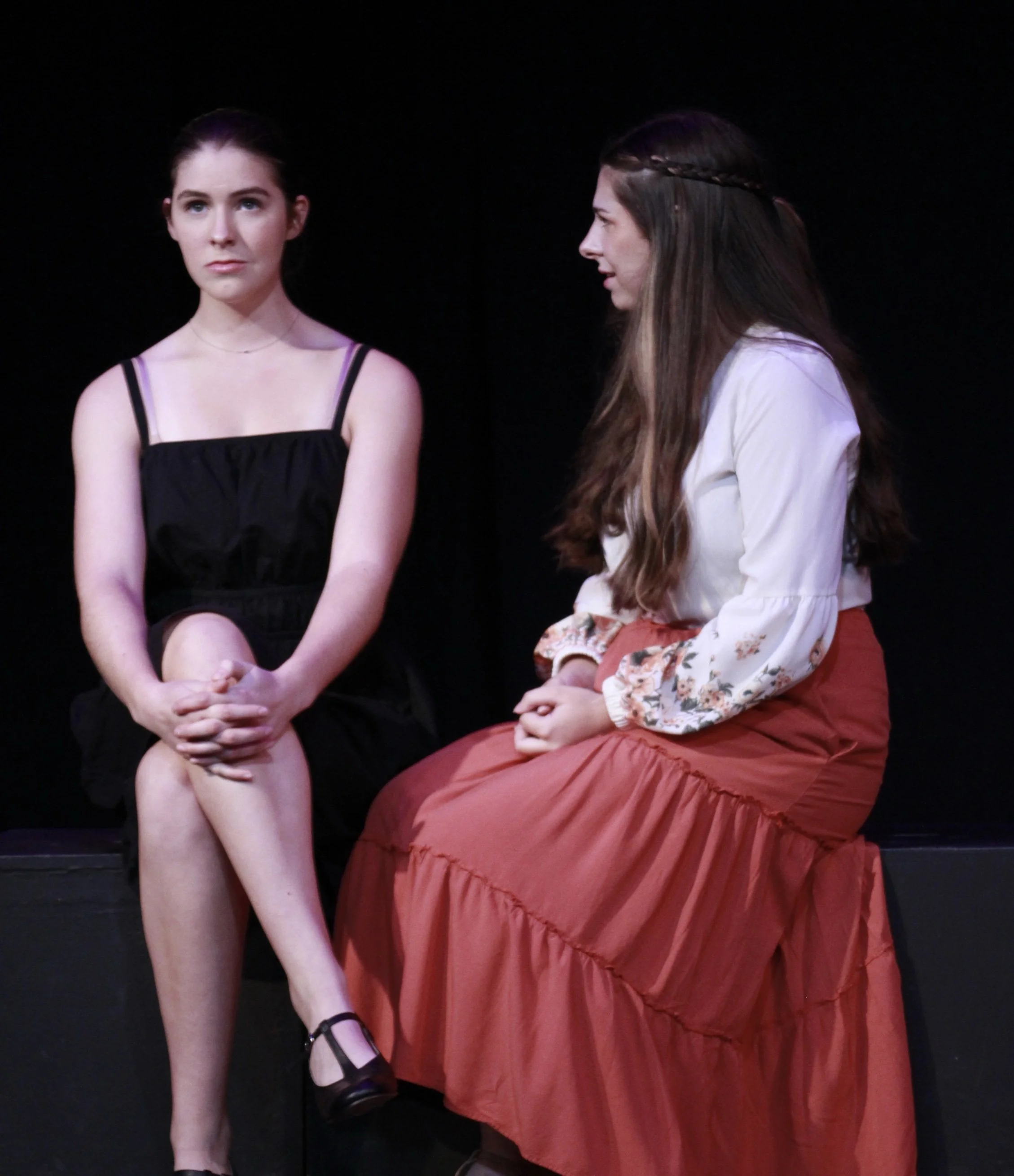 Two women sitting on a black stage, engaged in a conversation. One woman in a black dress with spaghetti straps, the other in a white blouse with floral sleeves and a red skirt.