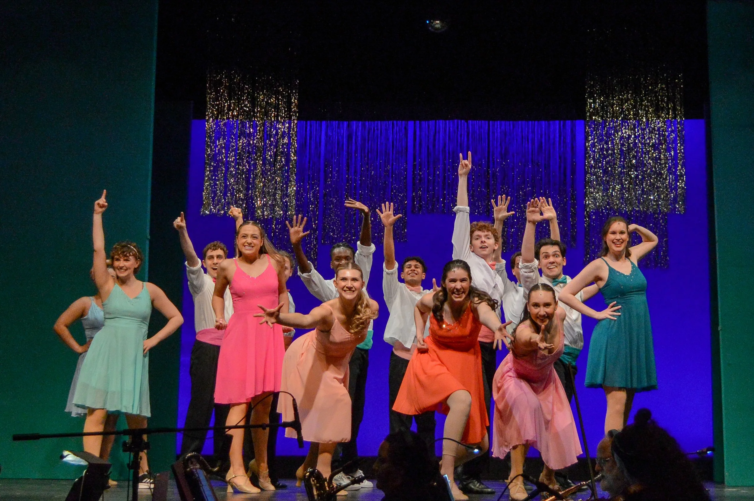 The cast of a musical theater performance on stage, with performers dressed in colorful costumes, smiling, and posing in front of a blue backdrop with shiny silver curtain decorations.