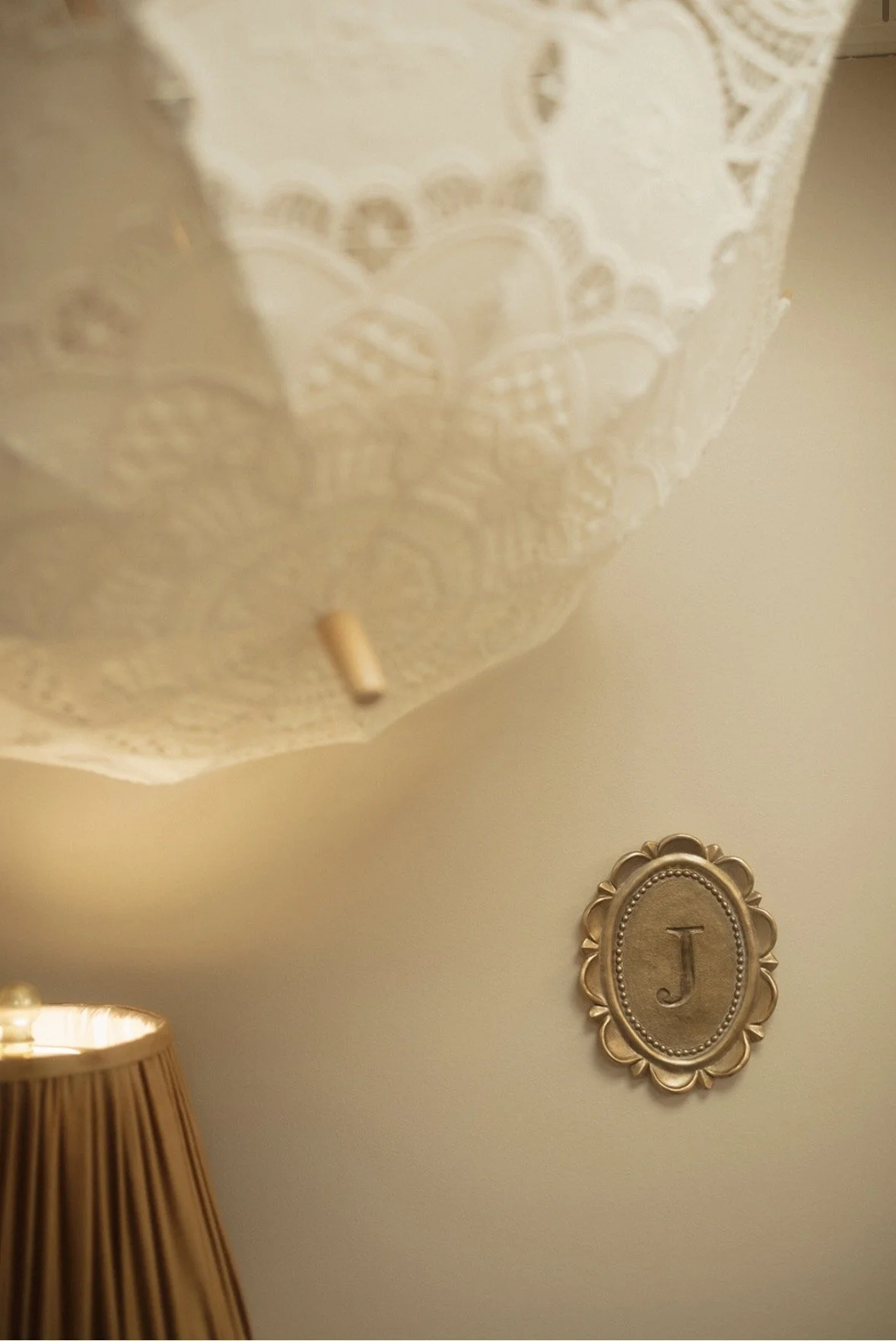A wall with an oval gold decorative frame with the letter 'J' inside, a beige lampshade, and a lace lampshade in the background.
