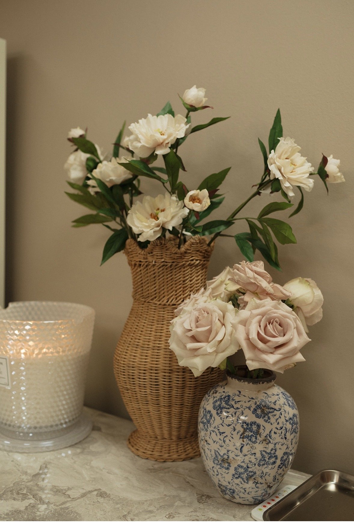 A marble countertop with two vases of flowers, one woven rattan and one blue and white ceramic, and a lit candle in a textured glass holder.