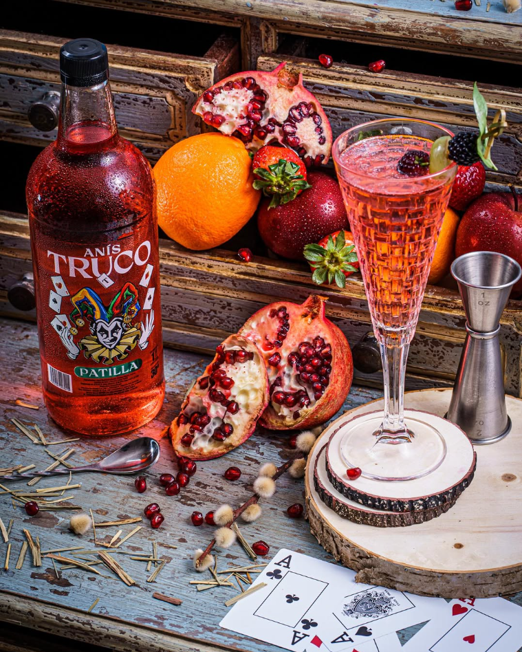A bottle of Anís Trucos Patilla on a rustic table, surrounded by pomegranate halves, strawberries, an orange, and a pink sparkling cocktail garnished with berries and mint. Playing cards and scattering of pomegranate seeds and dried herbs are also present.