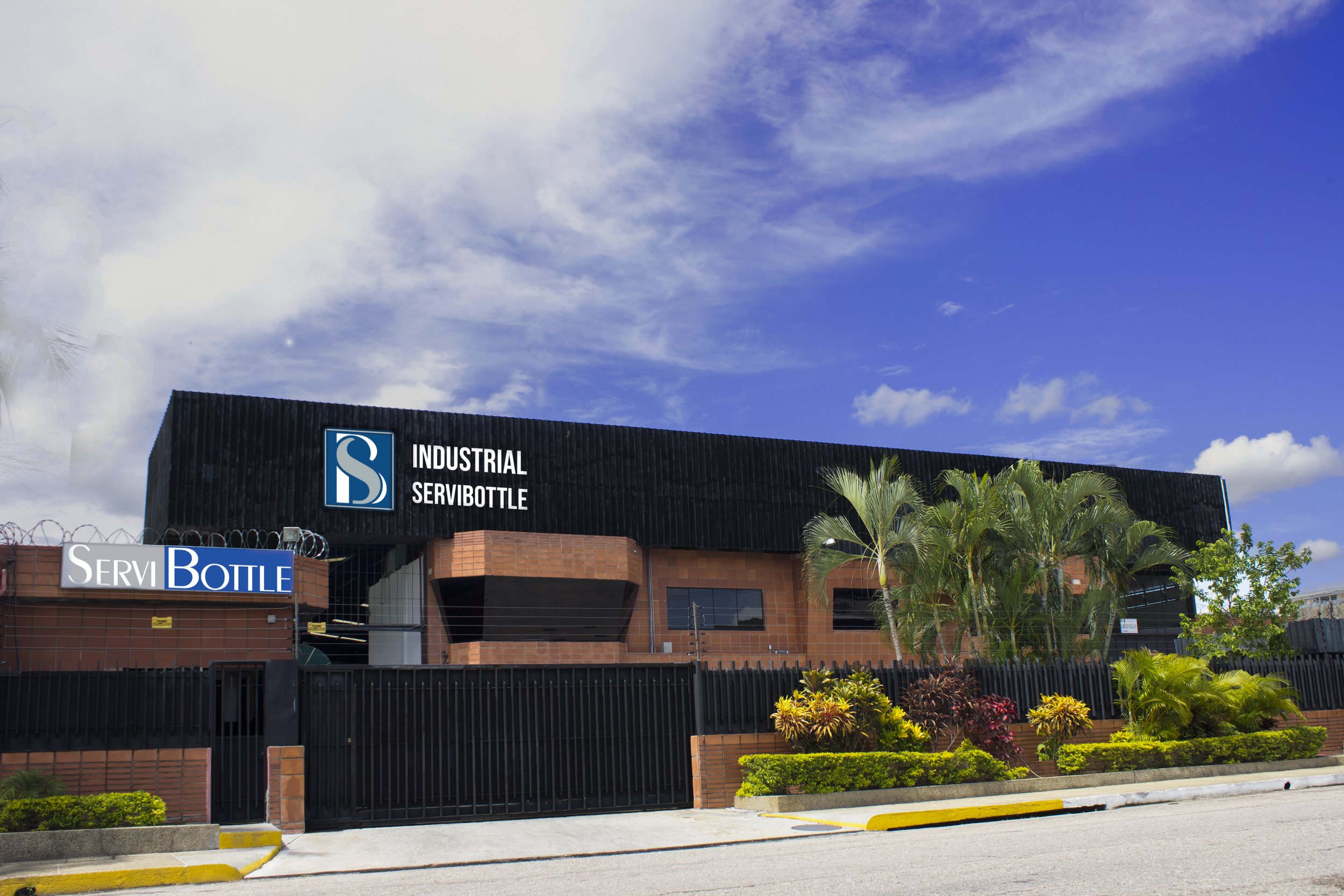 Industrial building with signs labeled 'Industrial Servibottle' and 'ServiBottle', surrounded by palm trees and plants under a partly cloudy sky.
