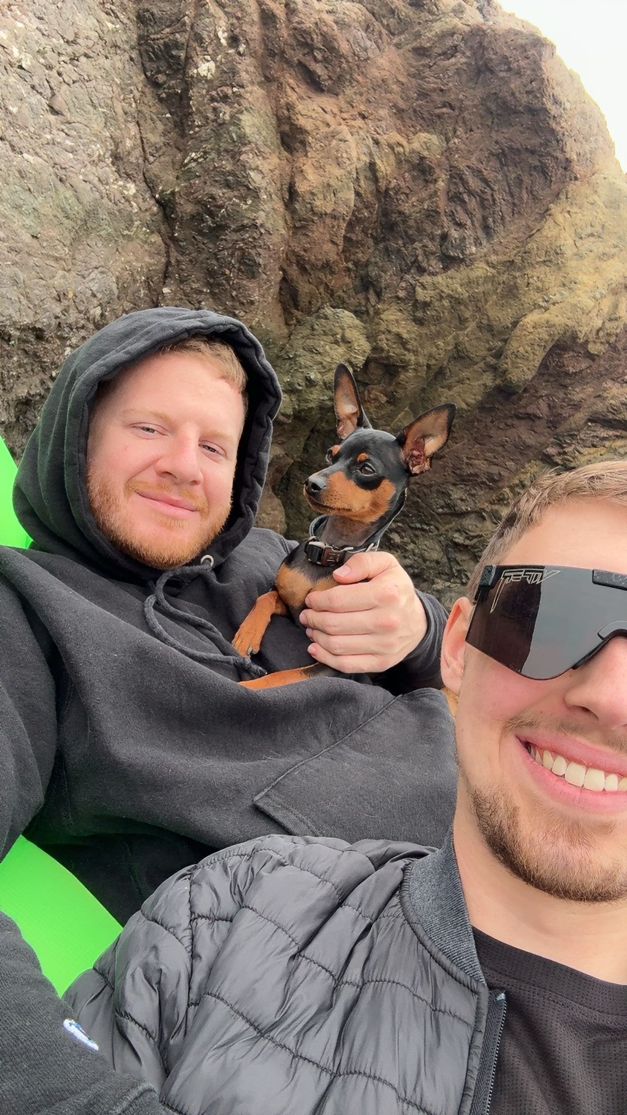 Two smiling young men taking a selfie outdoors with a small black and tan dog, against a rocky background.
