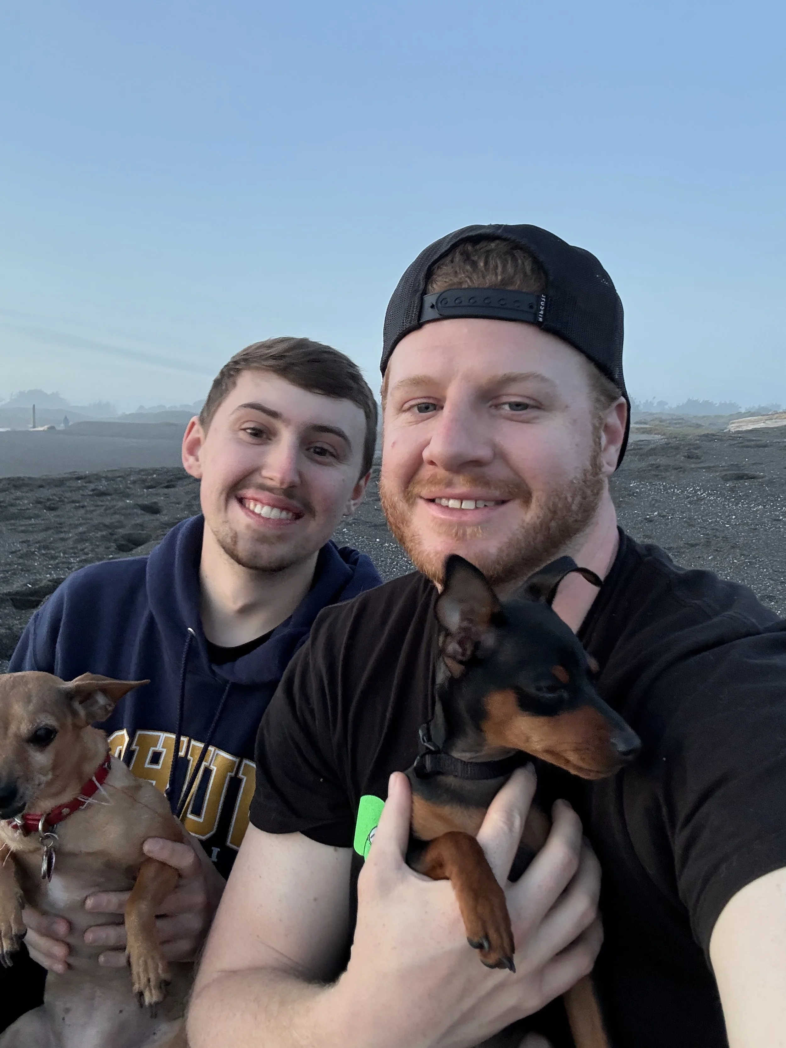 Two men smiling outdoors at dusk, each holding a small dog, with a rocky landscape and a light blue sky in the background.