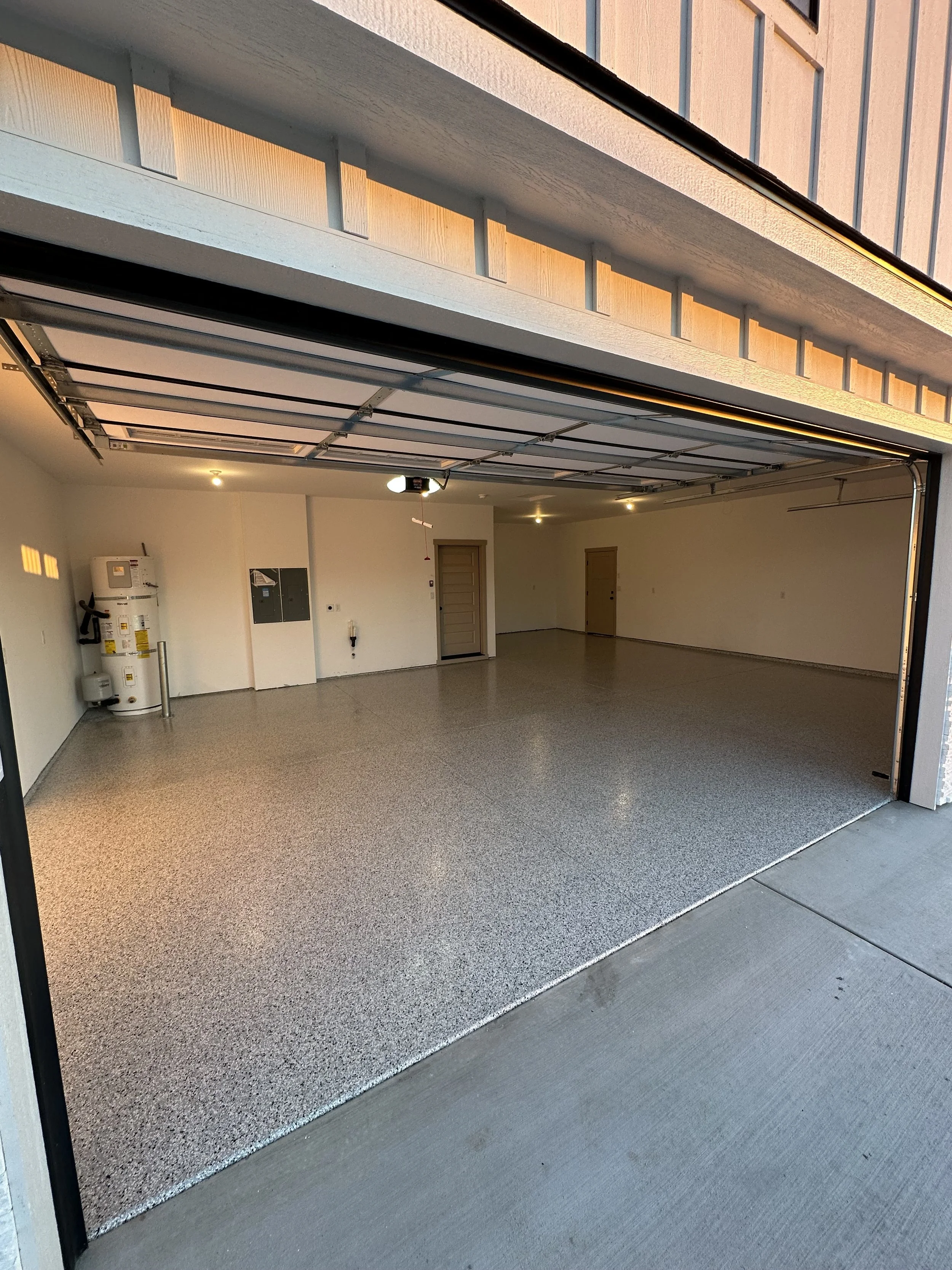 An empty residential garage with a newly finished epoxy floor, white walls, and a garage door open to outside. There is a water heater and electrical panels on the left wall, and two doors on the back wall.