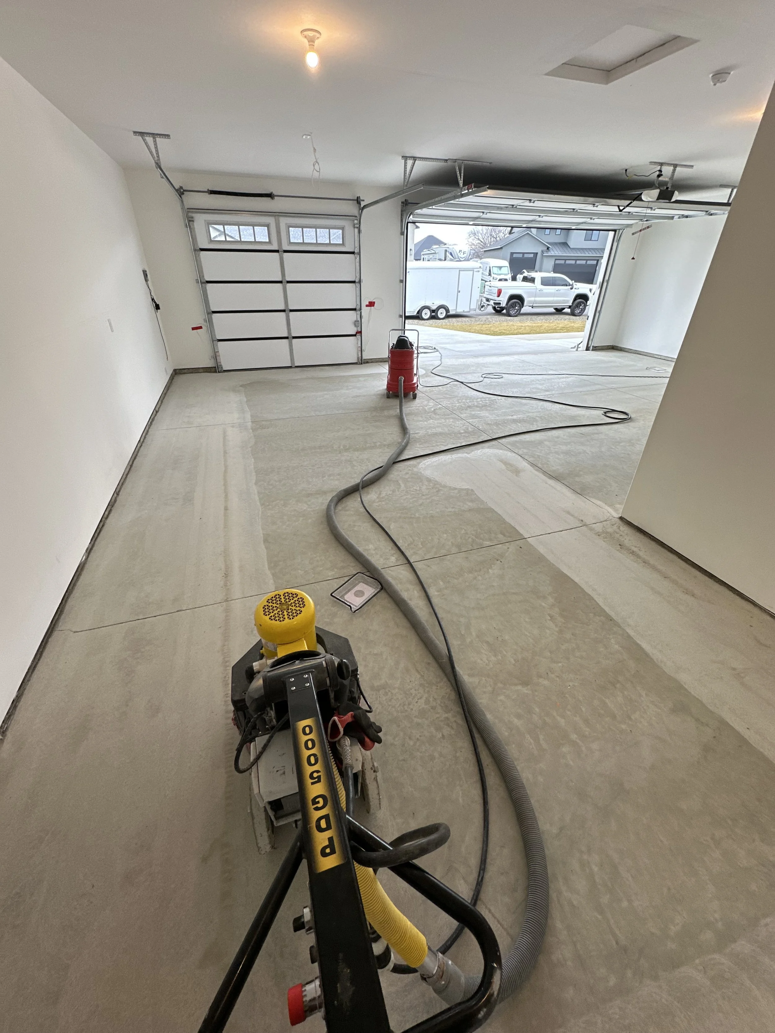 Interior view of a garage with a concrete floor being resurfaced, with tools and equipment, including a vacuum and hoses, and a partially open garage door showing parked cars outside.