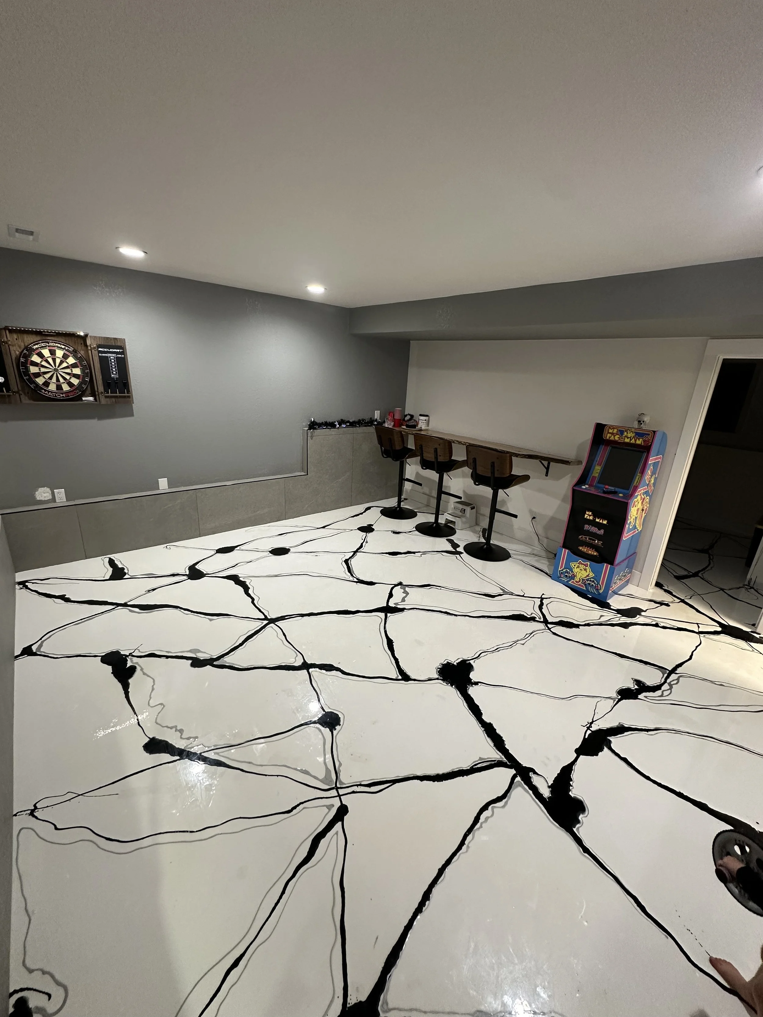 Basement with a large white floor featuring black abstract lines, a dartboard on the wall, a bar area with three stools, and a Pac-Man arcade game.