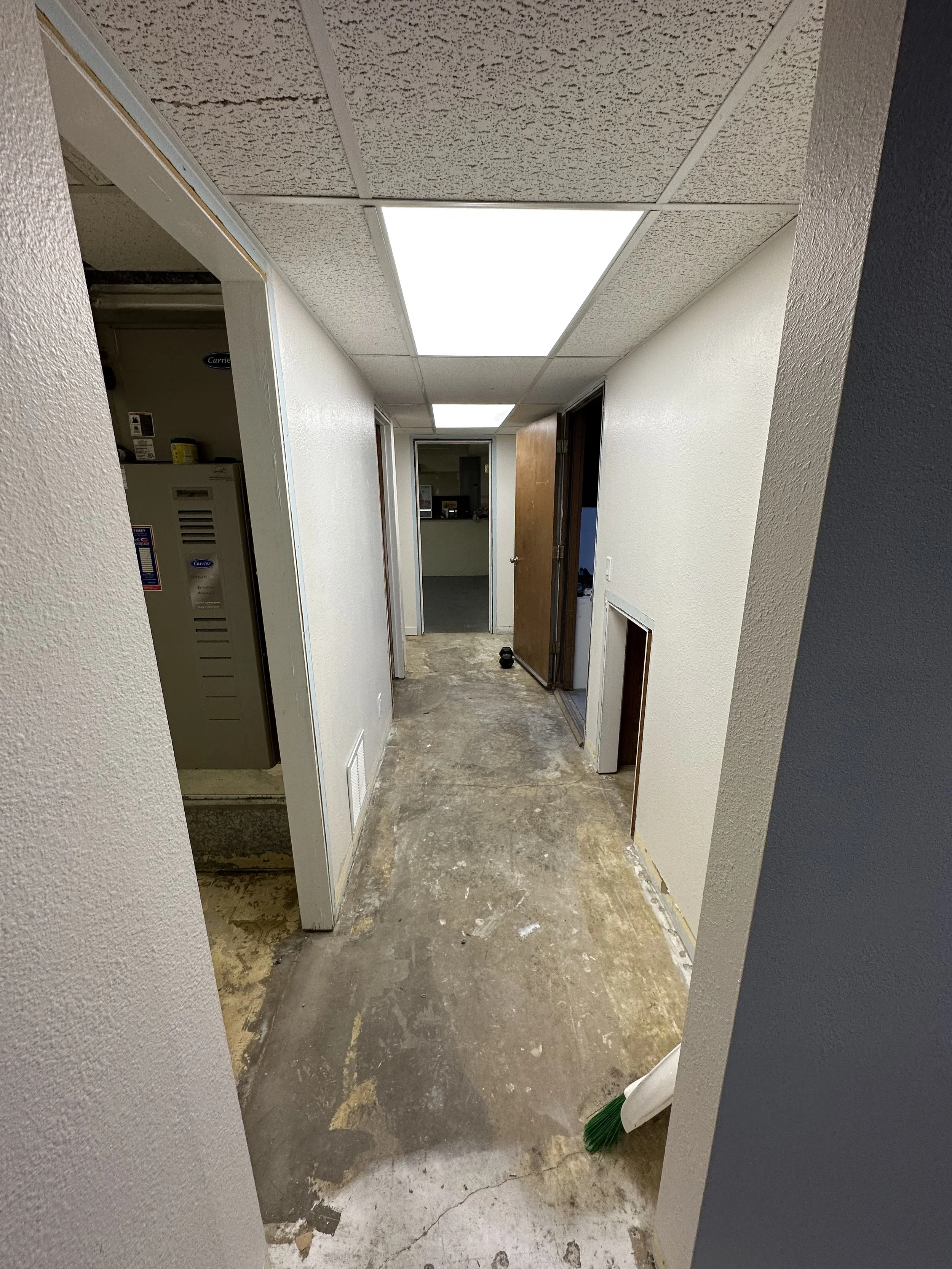 Interior hallway under renovation with exposed concrete flooring, white walls, a drop ceiling with fluorescent lights, and a partially open door on the right side. A broom is leaning against the wall on the right.