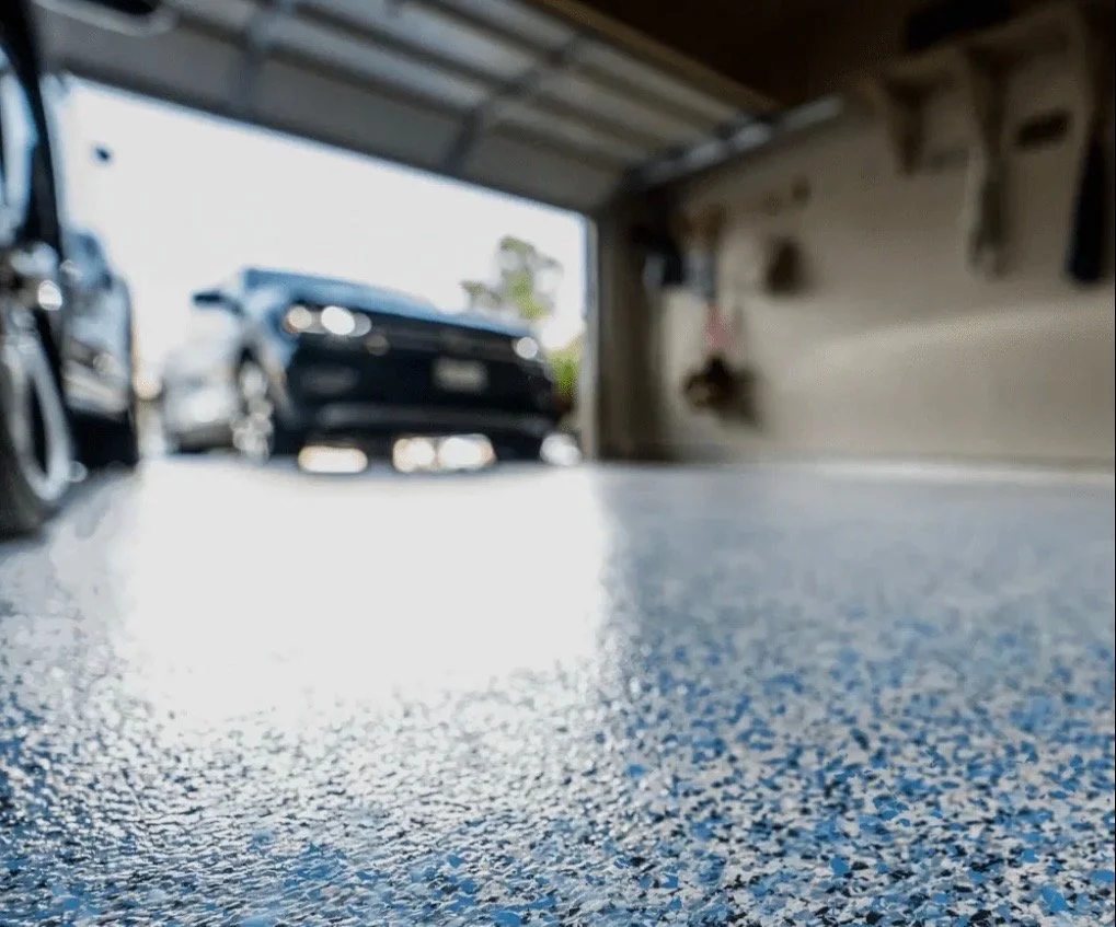 View of a garage with a textured floor, showing two cars parked outside and garage tools on the wall.