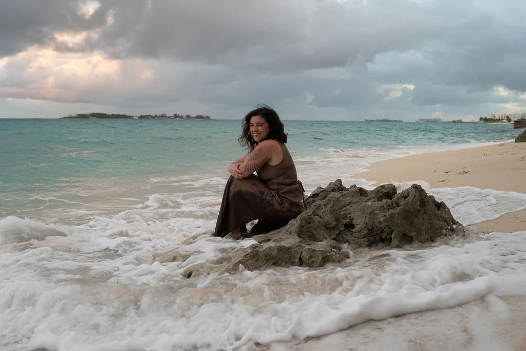 A somatic therapist sits on a rock on the shore of the sea.