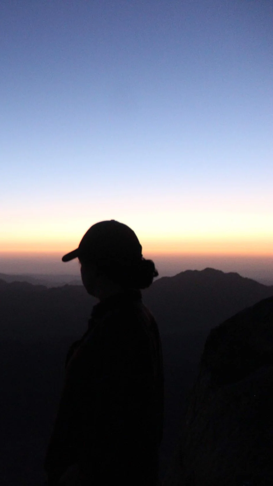 Silhouette of a person wearing a cap during sunset or sunrise on a mountain with a colorful sky and distant mountain ranges.