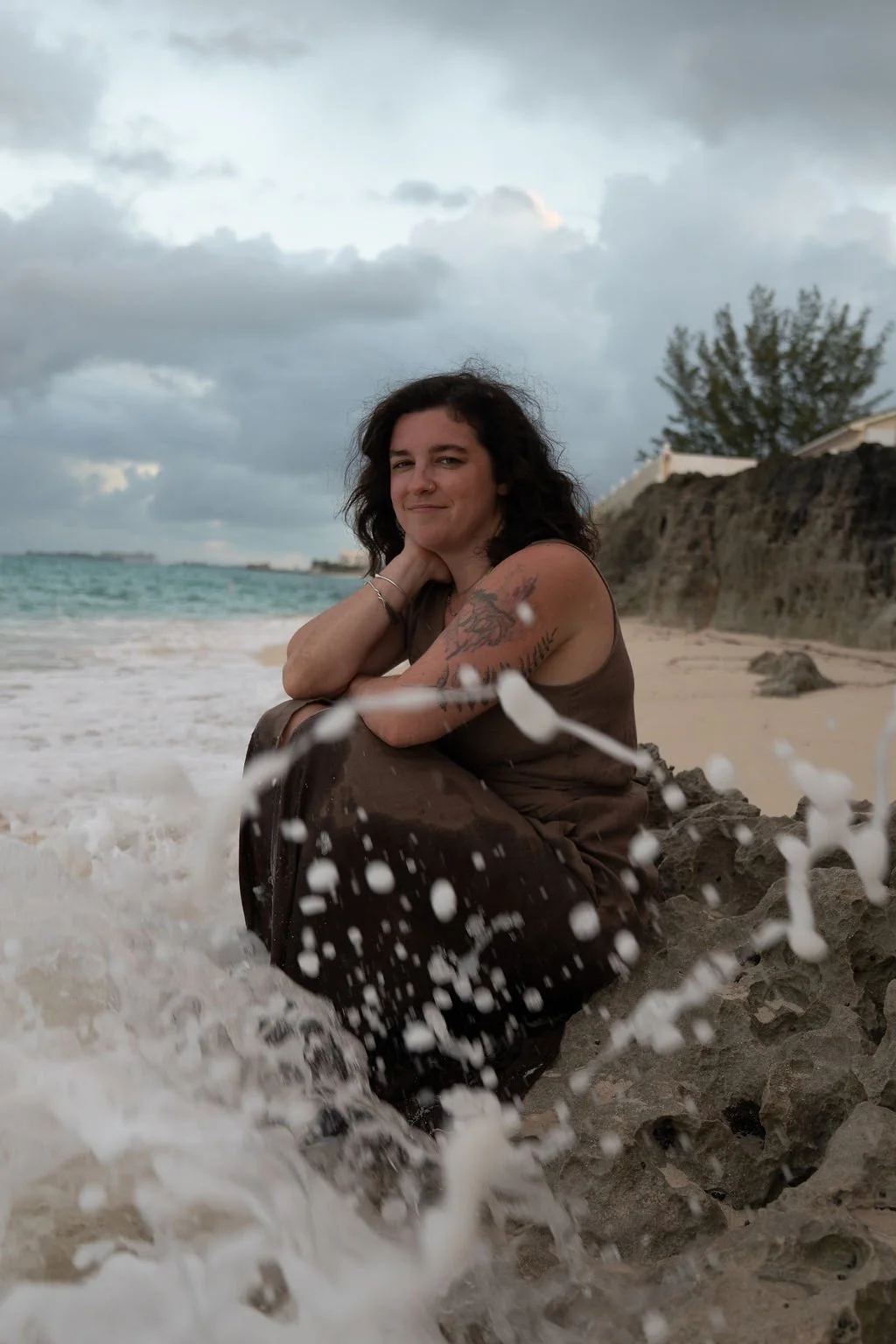Waves crash against a woman sitting on a rocky shore.