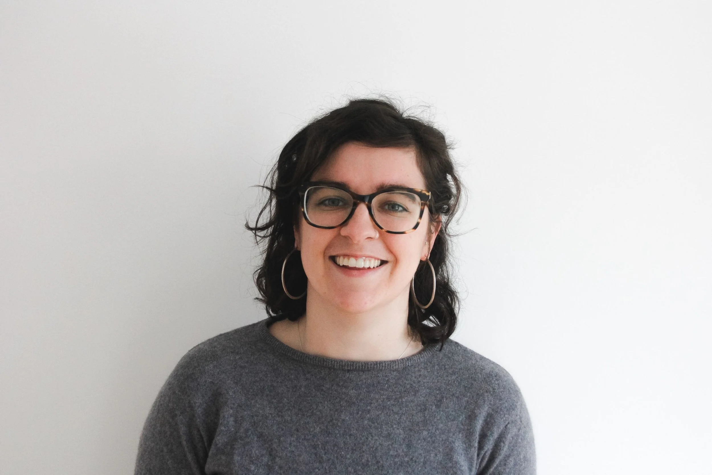 A woman with dark, wavy hair wearing glasses, earrings, and a gray sweater, smiling at the camera against a plain white background.
