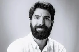 A smiling man with dark hair and a beard, indoors with large windows in the background.