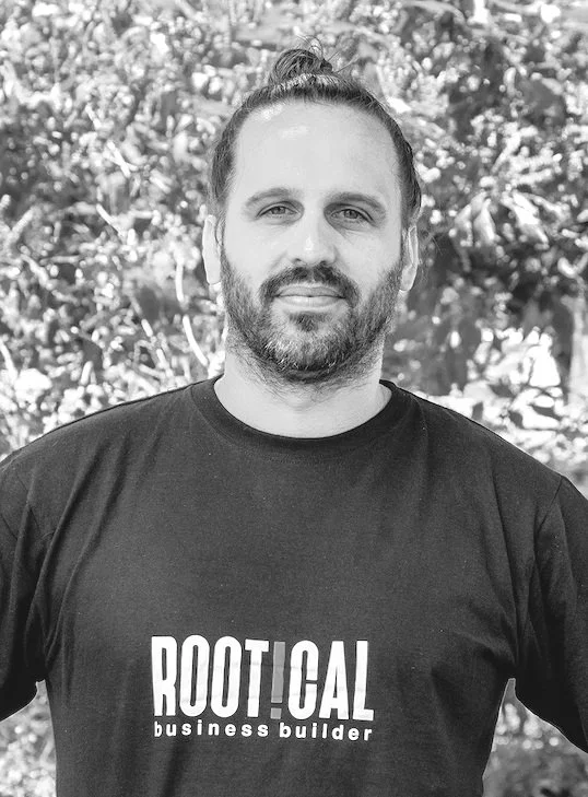 Man with beard and man bun smiling outdoors, wearing a black t-shirt that says "ROOTICAL business builder" in front of leafy trees.