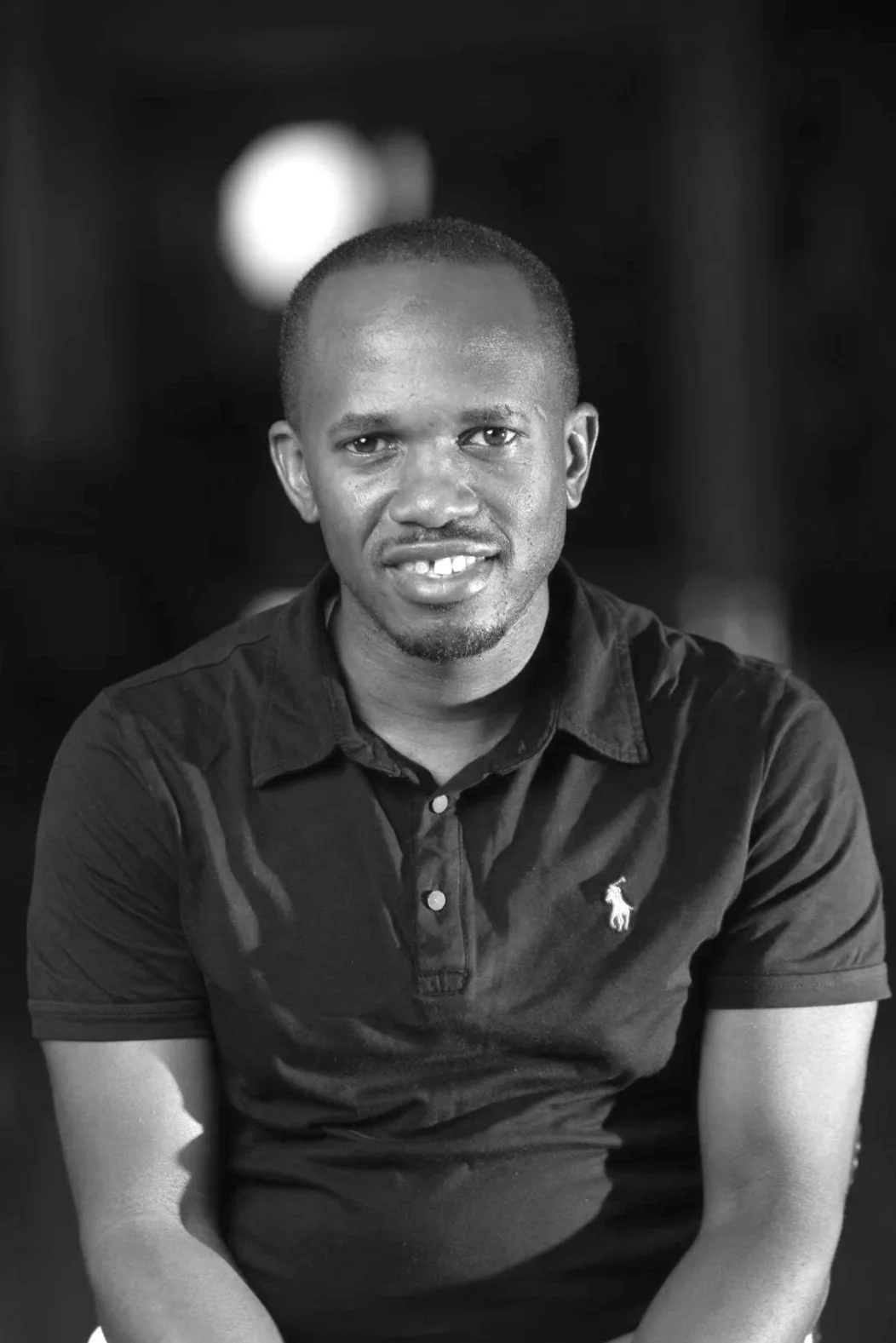 A young man with short hair, smiling slightly, wearing a black polo shirt with a small logo on the chest, sitting with arms resting on his lap against a dark background.