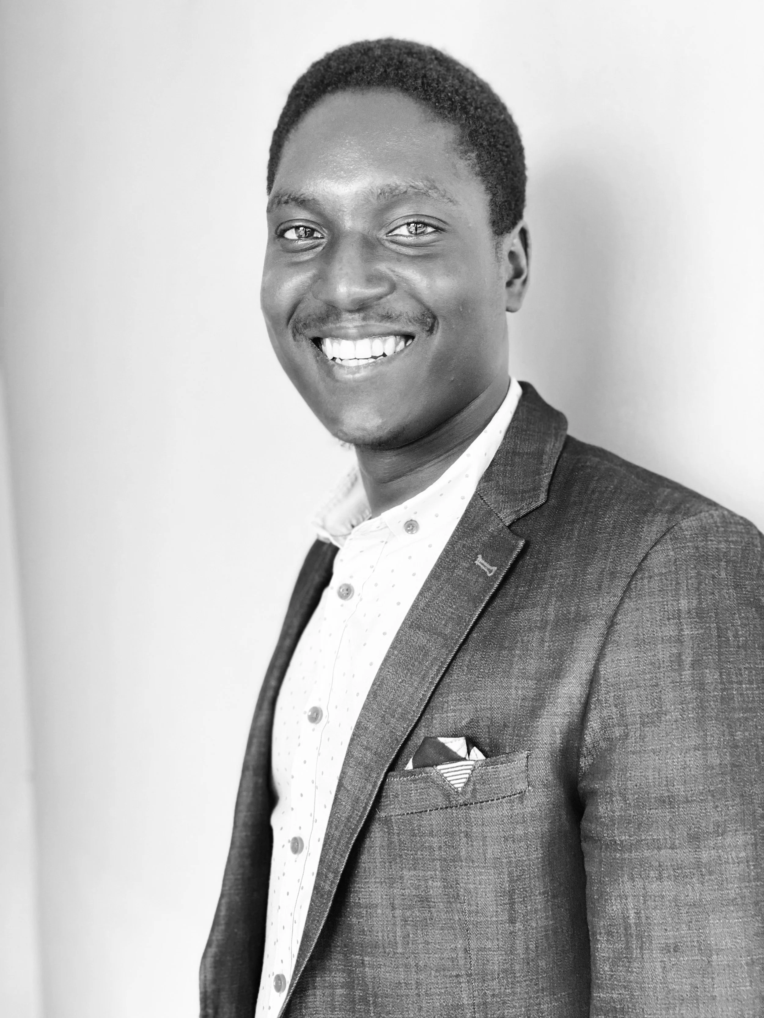 Black and white photo of a smiling young man wearing a blazer and patterned shirt, standing against a plain background.