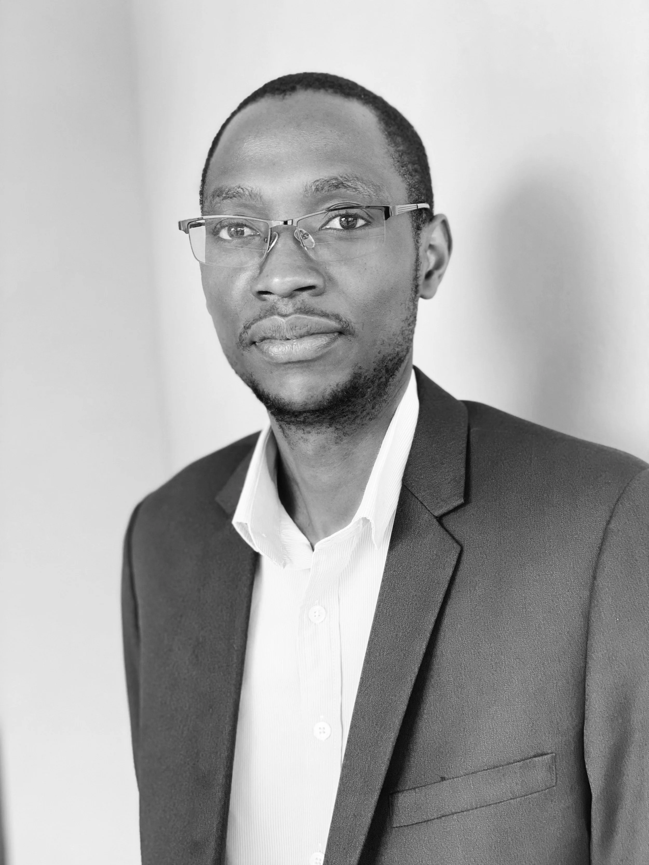 Black and white portrait of a man wearing glasses, a suit jacket, and a white collared shirt, standing against a plain background.